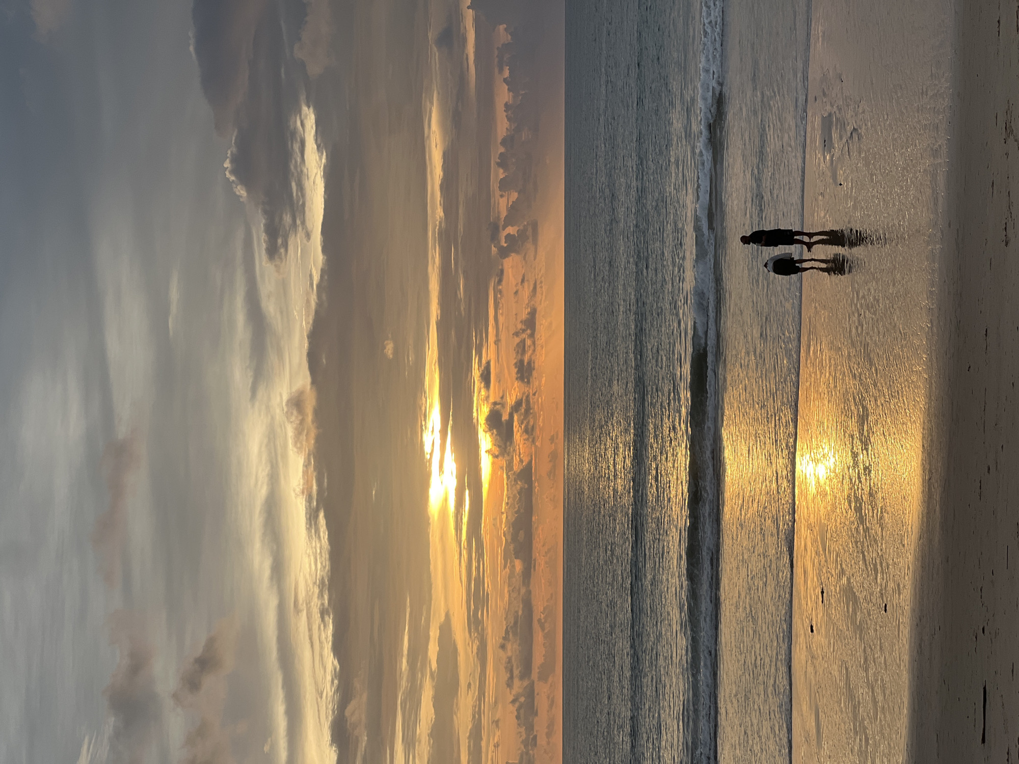 Two figures are silhouetted against the sunset on a beach in Choeng Thale, Thailand. The sky is filled with clouds tinged with orange and yellow from the setting sun, which is partially obscured by clouds. The ocean is a calm blue-gray, with gentle waves lapping at the shore. The wet sand on the beach reflects the vibrant colors of the sky, creating a mirror-like effect. One figure, appearing to be an adult, stands upright, while the other, possibly a child, is bent over, perhaps examining something on the sand. The figures are positioned near the water's edge. The overall scene evokes a peaceful and serene atmosphere.