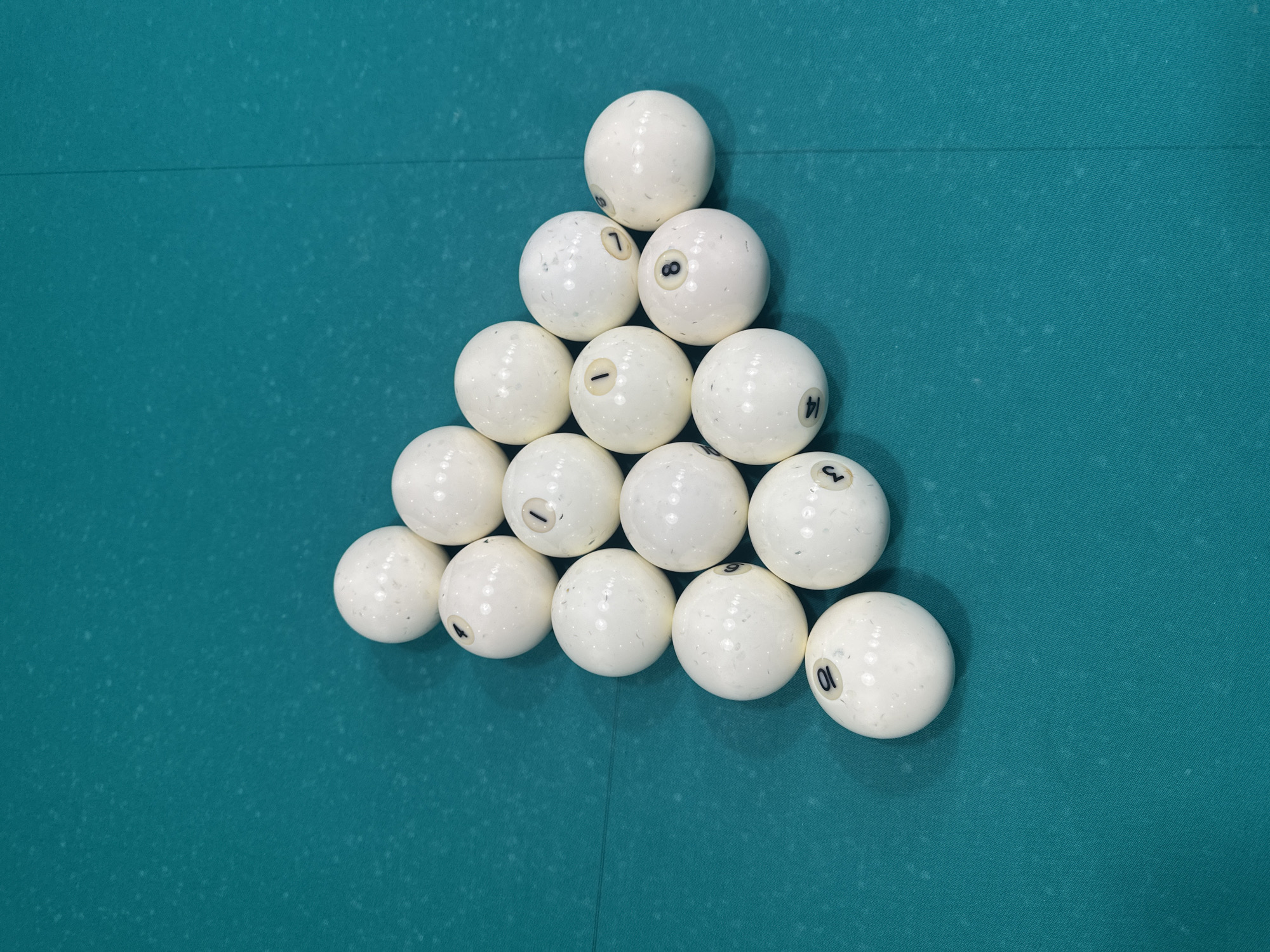 A rack of white billiard balls, numbered 1 through 10, is arranged in a triangular formation on a teal-colored felt surface, likely a billiard table. The balls are predominantly white with black numerals indicating their values. The balls are arranged in five rows, with the top row containing one ball, the second row two balls, the third row three balls, the fourth row four balls, and the bottom row five balls. The felt surface exhibits a subtle texture and a seam runs vertically down the right side of the image. There are no people or actions depicted in the image, nor is there any information or context to indicate the location in the city of Perm, Russia.