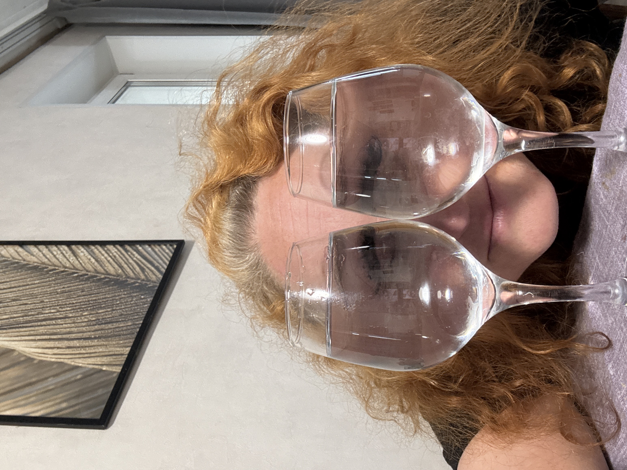An adult individual with long, wavy reddish-blonde hair, showing some grey at the temples, is the primary subject. Their face is partially obscured by two large, clear, stemmed glasses, each filled with a clear liquid (likely water), positioned over their eyes as if worn like spectacles. Their lips are visible, slightly parted.
The individual appears to be lying down, with their head resting on a light-colored, possibly lavender or light grey, textured fabric. The setting is indoors, likely a room in a home located in Chaikovsky, Russia. The background features a plain light grey wall. In the upper right background, a white-framed window or skylight is visible, allowing diffused natural light into the space. To the left of the person's head, a framed picture or artwork depicting a close-up of a feather or similar natural texture in neutral beige and black tones is mounted on the wall.
The activity captures a playful or artistic pose, with the glasses creating a surreal visual effect over the eyes. The diffused light suggests it is daytime. There is no visible text in the image.
