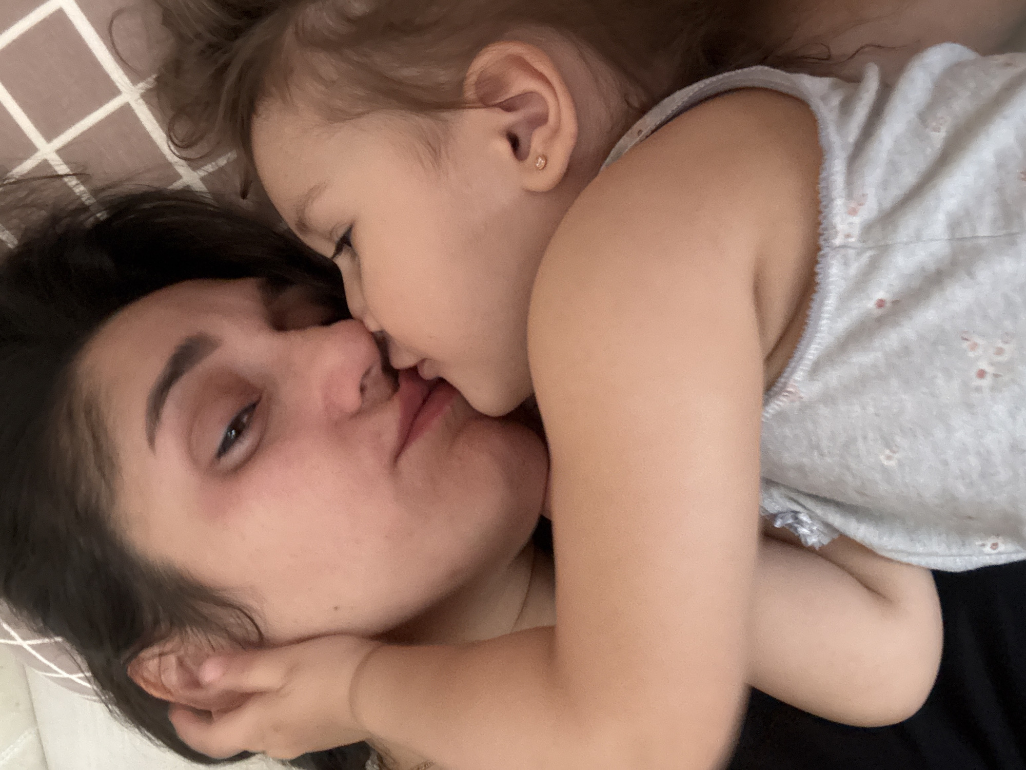 A close-up shot features a woman and a young child lying together on what appears to be a bed. The child is positioned on top of the woman, seemingly kissing her on the nose and lips. The woman has dark hair and a fair complexion, with her eyes slightly open. The child, also fair-skinned, has light brown hair and is wearing a light-colored top with small floral designs. The background includes a checkered pillow or blanket. The location cannot be determined from the image, and there is no indication of Tobolsk, Russia.