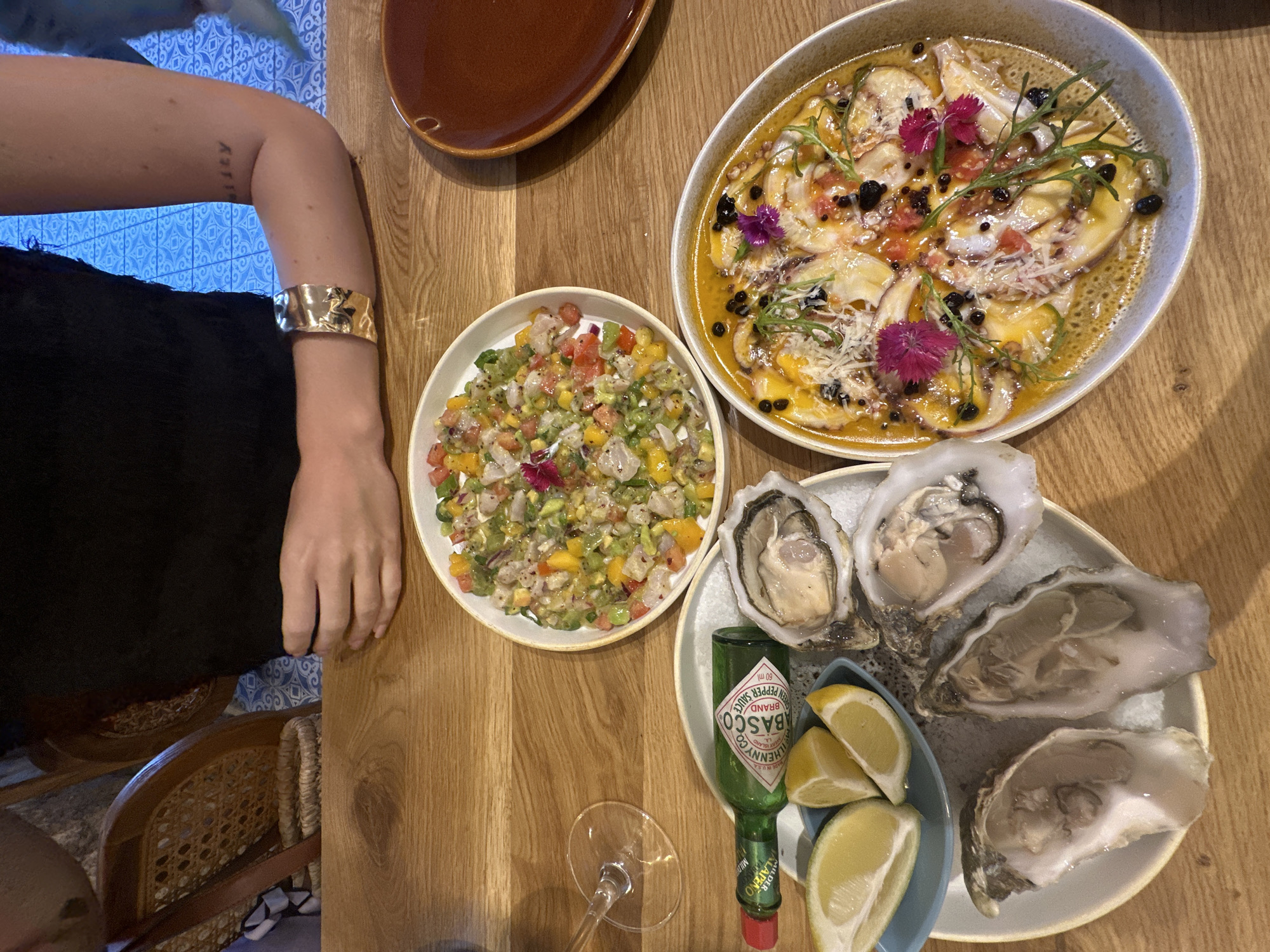 In Tel-Aviv, Israel, a person is seated at a wooden table, wearing a black top and a gold bracelet. Before them are three plates of food: one with oysters and lemon wedges, another with a colorful ceviche, and a third with a dish that appears to be octopus in a sauce, garnished with flowers and herbs. A bottle of Tabasco sauce is also visible. The background shows elements of a restaurant setting, including chairs and a tiled wall.