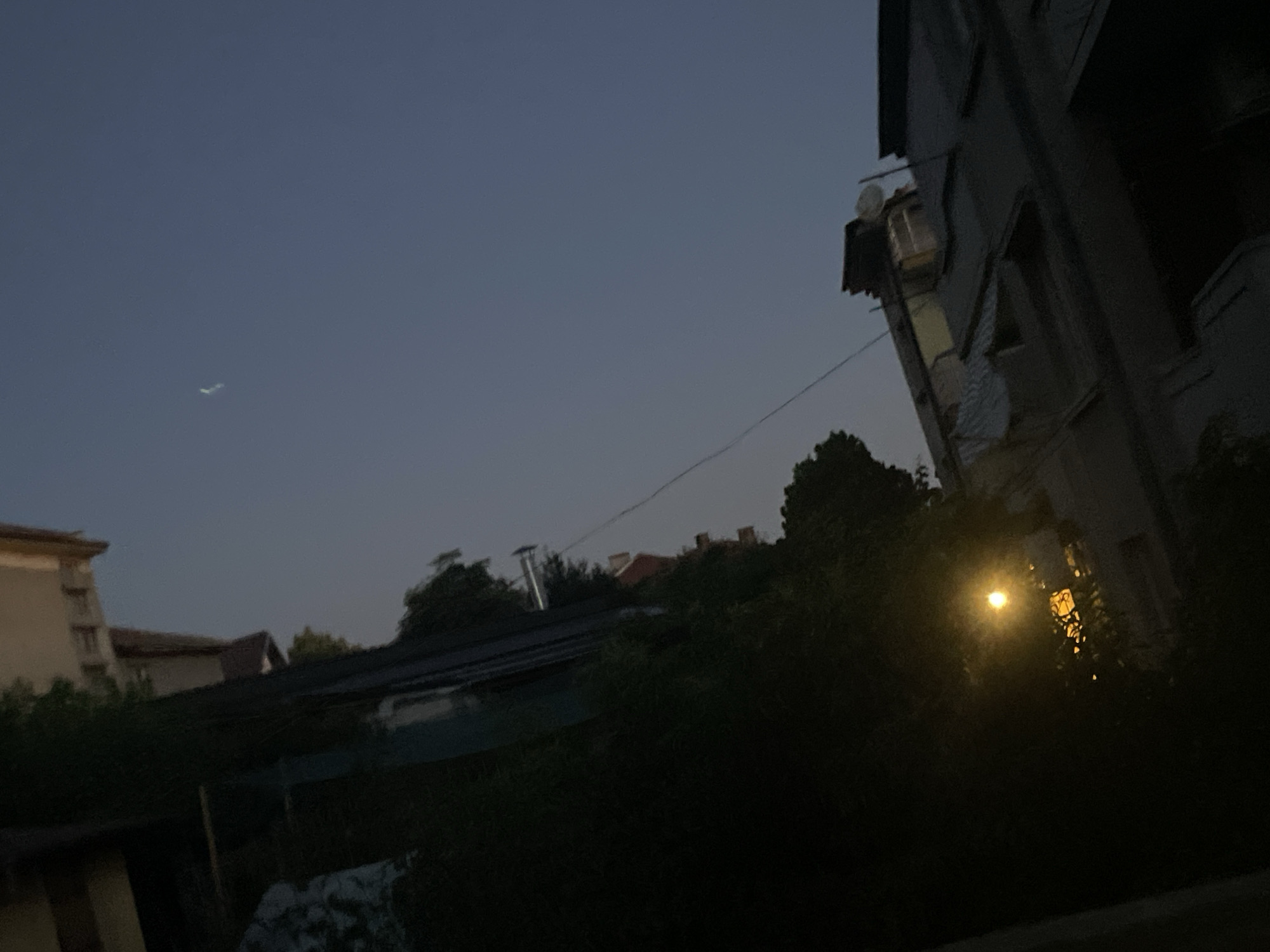 The image captures a twilight scene in Harmanli, Bulgaria, with a clear, darkening sky. A faint contrail from an aircraft is visible in the upper left quadrant. In the foreground, the silhouette of dense vegetation is present, obscuring much of the ground level. To the right, the corner of a multi-story building is visible, with a bright, illuminated window or light source emanating from within, casting a warm glow. The architectural style of the building appears to be somewhat dated. Further to the left, the edge of another building is partially visible, suggesting an urban or suburban residential area. The overall impression is one of a quiet evening in Harmanli, with hints of both human activity and the natural environment.
