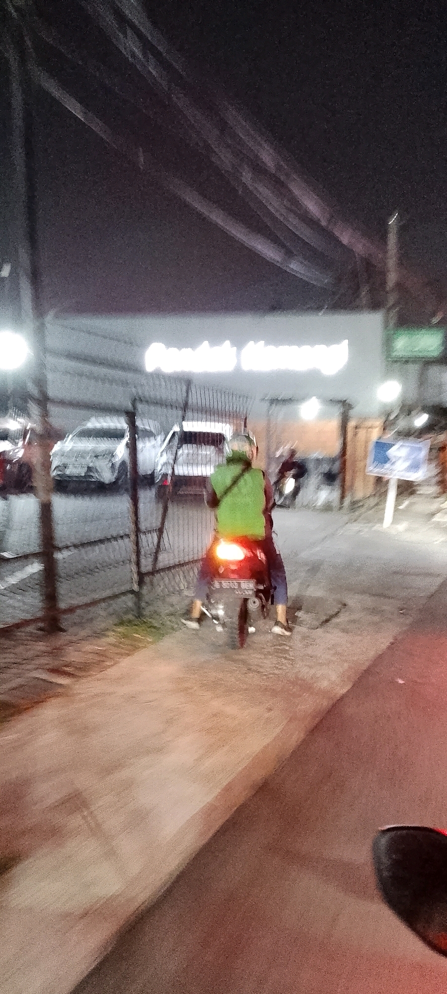 A low-angle, slightly blurred nighttime photograph captures a street scene in Gunung Putri, Indonesia. The foreground is dominated by a reddish-brown paved area and a dark, partially visible object, possibly the handlebars of a motorcycle. In the mid-ground, a person on a motorcycle is seen from behind. The rider wears a bright green vest and dark pants, and a helmet. The motorcycle's taillight is illuminated. Behind the motorcycle, a metal fence separates the road from a parking area. Several cars are parked behind the fence, their bodies appearing to be covered in white material, perhaps protective wraps. A brightly lit sign with text, possibly advertising, is visible on a pole to the right. In the background, a building with a large, illuminated sign reads "Domino's Pizza". Overhead, power lines crisscross the dark sky. The lighting suggests artificial illumination from streetlights and the building's signage.