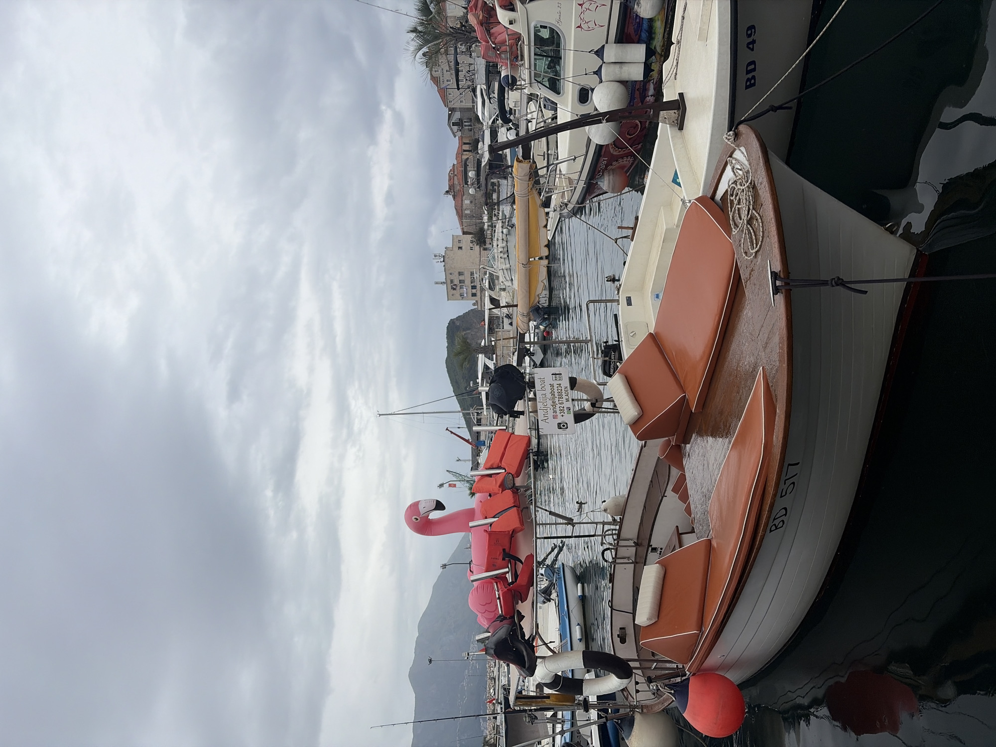 The photograph depicts a harbor scene in Budva, Montenegro. The sky is overcast with heavy clouds. Several boats are moored in the harbor. A small boat with orange cushions and the lettering "BD 517" is prominent in the foreground. It is secured next to a larger boat labeled "BD 49." A bright pink flamingo float is visible on another boat alongside several orange life jackets. The harbor water is calm, with reflections of the boats and surrounding structures. Buildings and mountains can be seen in the background. Other boats and their rigging are visible in the midground.