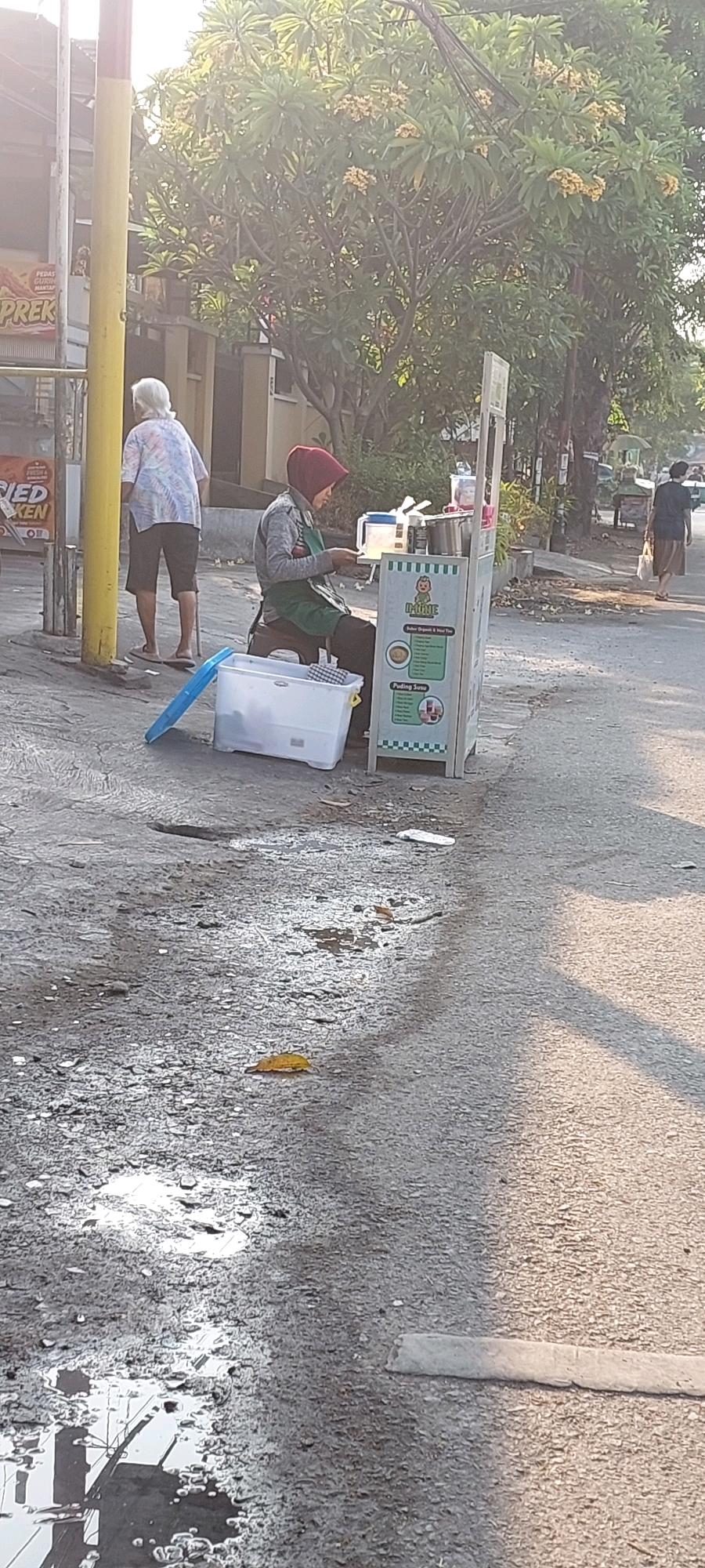 The image depicts a street scene in Semarang, Indonesia, likely in a commercial area given the presence of various food vendors. The foreground is dominated by a wet asphalt road with puddles reflecting the surrounding environment, including what appears to be a street light pole. In the mid-ground, a woman wearing a red hijab and a green apron is seated on a stool behind a mobile food cart. The cart has a sign with Indonesian text and graphics, suggesting it sells beverages or desserts. To the left of the cart, a large white plastic container with a blue lid is present. Further left, an elderly person with white hair, wearing a patterned shirt and shorts, stands near a yellow pole. The background shows trees with yellow flowers, residential buildings, and other street elements, including another food cart and a person walking away in the distance. The lighting suggests it is either early morning or late afternoon.