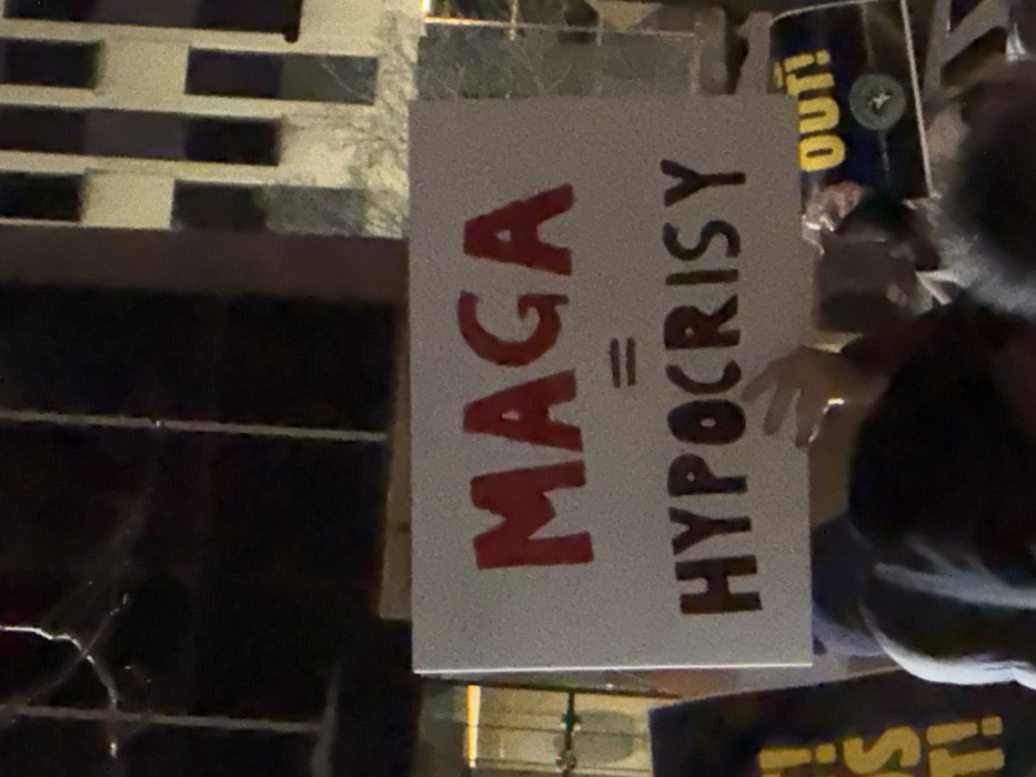 A protest sign reads "MAGA = HYPOCRISY" in red and black marker. The sign is held by a person with visible hands. A building with a black and gray grid pattern is in the background. To the right, another sign reads "OUT!" in yellow. The scene appears to be outdoors at night.