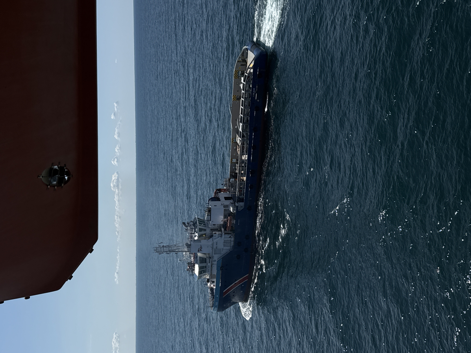 This aerial image, oriented vertically, shows a dark blue vessel with a white superstructure navigating through the ocean. The ship is leaving a white wake behind it, indicating movement. The sea is a deep blue, with visible ripples and some whitecaps, suggesting a moderate sea state. In the foreground, a large, reddish-brown object, possibly part of another vessel or structure, partially obscures the left side of the image. A circular, metallic fixture with grates is visible on this foreground object.
The sky appears light blue with some scattered white clouds. The sun is bright, casting reflections on the water's surface. The overall lighting suggests it is daytime, likely midday or early afternoon.
On the side of the dark blue vessel, some text is visible in white lettering. The words "STENA" and "BLUE" are discernable. The vessel appears to be a working ship, possibly a supply or support vessel, with railings and various equipment visible on its deck.
The scene is one of maritime activity in an open ocean environment. The image is taken from a high vantage point, looking down on the ship and the water. There are no visible people. The mood conveyed is one of industrial operation at sea.