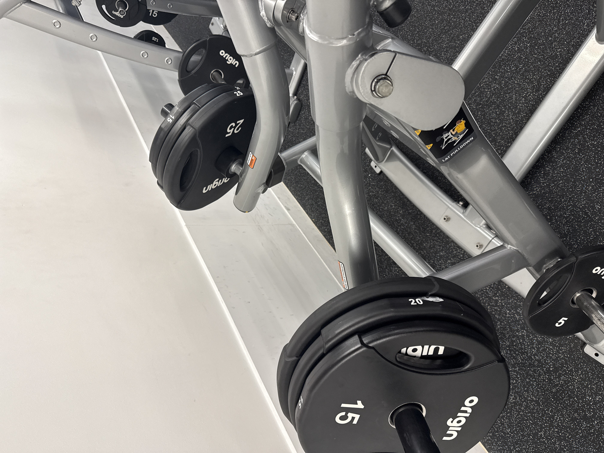The image presents a close-up view of gym equipment, specifically black weight plates and parts of a metallic exercise machine, situated in an indoor gym environment. No people are visible in the scene. The setting is identified as a gym in London, United Kingdom.
In the foreground and central area, several black 'origin' brand weight plates are stacked on a grey metal bar, which is part of a larger machine. Clearly visible numbers on these plates include '15' (twice) and '20'. To the left, another set of two 'origin' plates are stacked, both marked '25'. Further in the background, another 'origin' plate shows '15'. On the far right, a small black plate marked '5' is partially visible.
These weights are mounted on a robust, grey metallic gym machine frame, constructed from thick, tubular components. An instructional sticker is affixed to one of the machine's beams, featuring an illustration of an exercise and the clear text "LAT PULLDOWN". The floor is a dark grey, speckled gym mat, providing a typical gym surface. A bright white wall or partition runs along the left edge of the image. The picture is taken at a tilted angle, giving a rotated perspective of the equipment. The lighting appears even, consistent with an indoor setting.