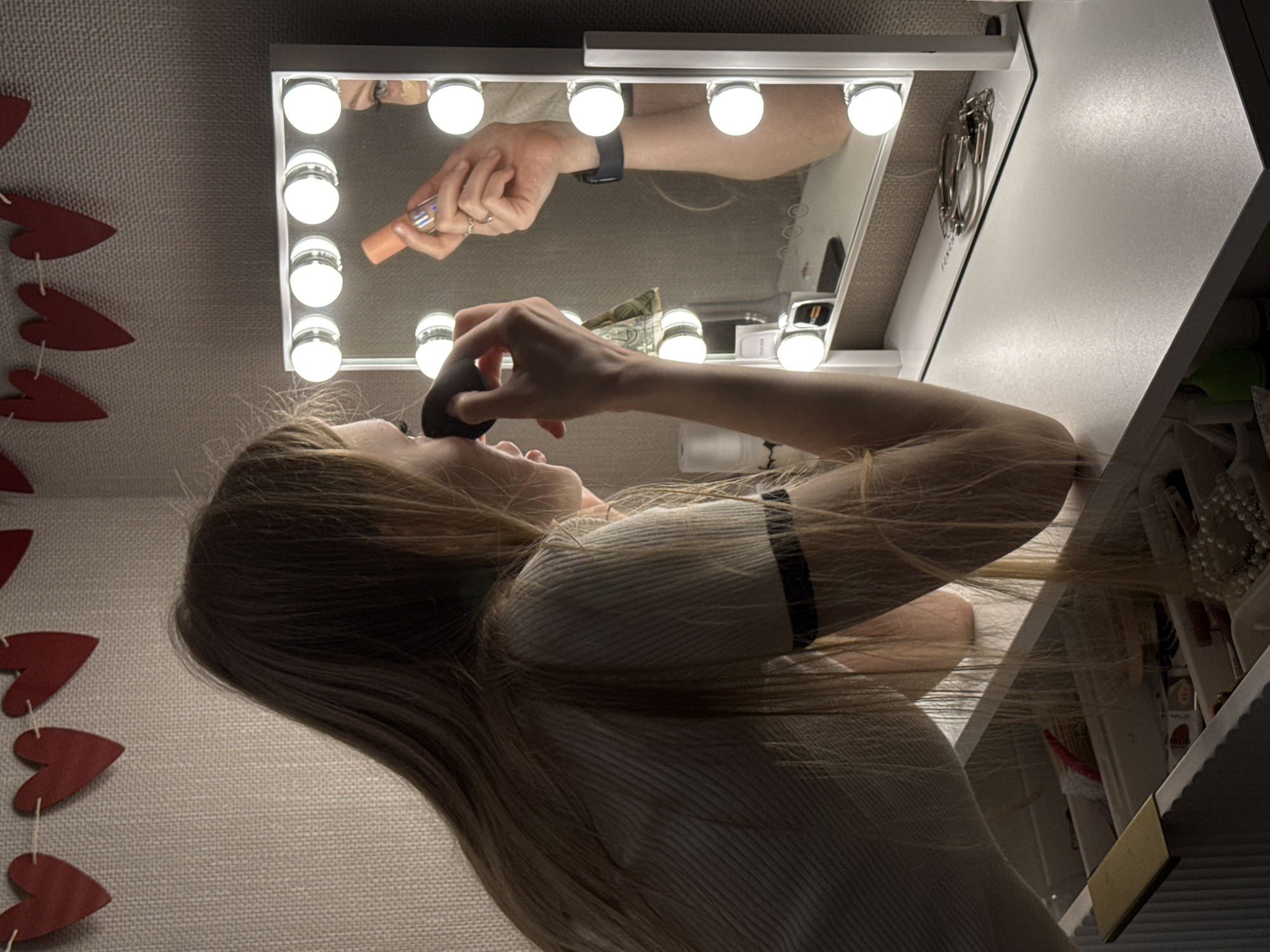 A woman with long blonde hair applies makeup in front of a lighted vanity mirror. She holds a makeup sponge near her face. The reflection in the mirror shows her other hand holding a tube of makeup. The background includes a wall decorated with red heart shapes. A vanity cabinet with various items inside is partially visible. The location and city of Perm, Russia are not discernible from this image.