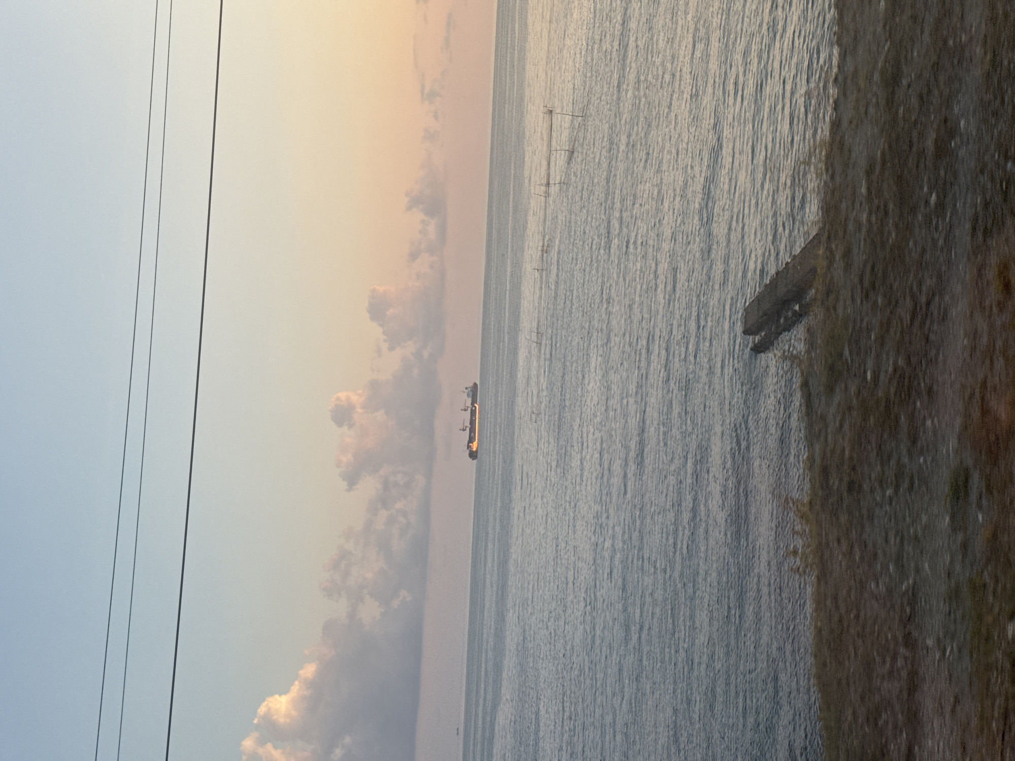 The image depicts a tranquil seascape at dusk, likely near the coastal town of Shepsi, Russia. Three thin power lines are visible in the upper portion of the frame, stretching horizontally across the sky. Below them, a soft gradient of colors paints the sky, transitioning from a pale blue at the top to a warm, peachy hue near the horizon. Fluffy, scattered clouds, tinged with pink and orange light, drift across the sky.
The vast expanse of the sea dominates the mid-ground. Its surface is calm, with gentle ripples reflecting the muted light. In the distance, a large cargo ship is visible, its silhouette dark against the horizon, with some yellow lights illuminating its structure. Closer to the shore, a series of thin poles emerge from the water, possibly part of a fishing net or a marker system.
The foreground consists of a rough, uneven shoreline covered with dry grass and small stones. A weathered, concrete structure, possibly a section of a pier or seawall, juts out from the bank into the water. The overall atmosphere is serene and still, capturing a quiet moment by the Black Sea.