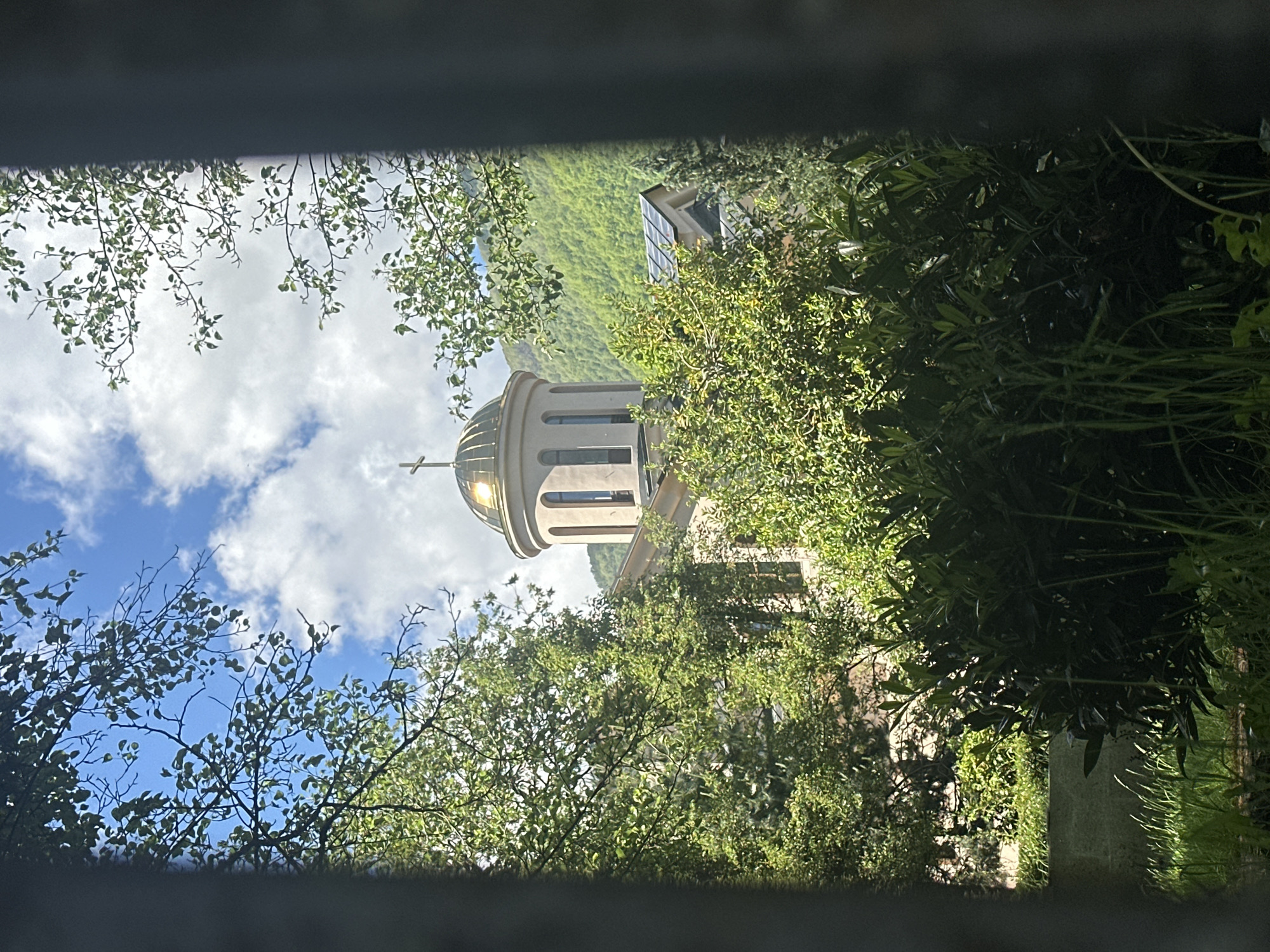 A vertically oriented outdoor photograph, framed through a narrow, dark opening, captures a scenic view centered around a domed building nestled amidst lush greenery under a bright sky. The dark, linear bands at the top and bottom suggest the view is through a slit, perhaps a window or fence.
The main subject is a light-colored building, likely a chapel or church, featuring a prominent, rounded dome topped with a cross. The dome has a polished, reflective surface, possibly metallic, which catches the sunlight. Several arched windows or decorative recesses are visible on the cylindrical base of the dome. This structure is partially enveloped by dense foliage and trees with vibrant green leaves, which occupy the foreground and midground, casting dappled shadows.
In the background, a verdant mountain slope, completely covered in trees and vegetation, rises towards the horizon. A section of another, more modern-looking building with a sloped roof and reflective panels is partially visible on the upper right side, blending into the mountain backdrop. The sky above is a clear, brilliant blue with scattered, fluffy white clouds.
The scene is bathed in bright daylight, indicating either morning or early afternoon, and the weather appears sunny and pleasant. The overall impression is one of tranquility and natural beauty, consistent with a mountainous, forested region like Krasnaya Polyana, Russia. There is no human activity or visible text in the image.