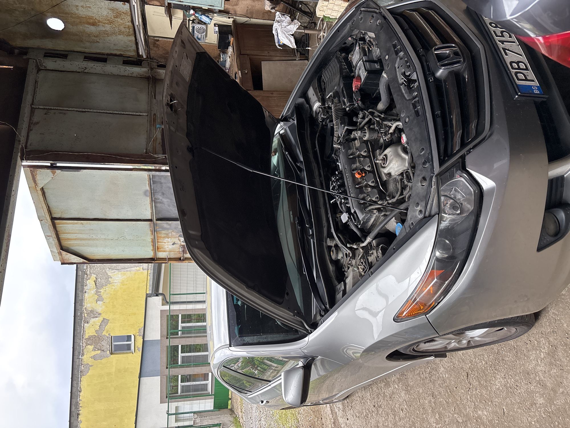 A silver Honda car is parked outdoors with its hood fully open, exposing the engine bay. The front of the car displays a Bulgarian license plate, "PB 7158 BG," indicating its registration in the Plovdiv region. The Honda emblem is visible on the grille, and "HONDA" is also legible on the engine cover.
The setting appears to be an older, possibly industrial or semi-urban environment in Zlatograd, Bulgaria, under an overcast sky. To the immediate left of the car (in the image's original orientation), there's a rustic metal structure with a rusty ceiling featuring a circular opening and various cables strung about. Further back in this area, shelves and scattered objects suggest a workshop or storage space. To the right, an old building with significant peeling yellow paint and a single visible window stands, next to a more modern structure featuring multiple windows, balconies, and green blinds.
No individuals are present, but the open hood indicates that the car is either undergoing maintenance, inspection, or has recently been worked on. The ground beneath the car is a concrete or asphalt surface. The overall scene conveys a functional and somewhat aged atmosphere.