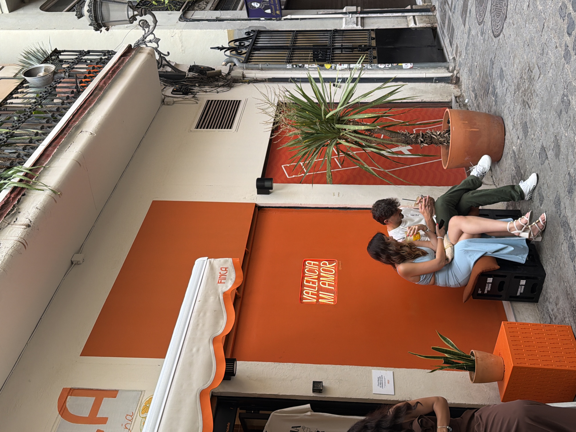 This is a medium shot of a street scene in front of an orange-colored building. Two people, a woman and a man, are sitting on black crates in front of the building. The woman, wearing a light blue dress, is looking down at her phone. The man, wearing a white t-shirt and green pants, is also looking at his phone and holding a drink. To the left of the couple, there is an orange decorative cube with a potted plant on top. A large potted palm tree stands to the right of the couple. Above them, a balcony with an ornate railing overlooks the street. The street itself is paved with cobblestones. The building has a white awning with orange trim. A neon sign that reads "VALENCIA MI AMOR" is visible on the orange wall.