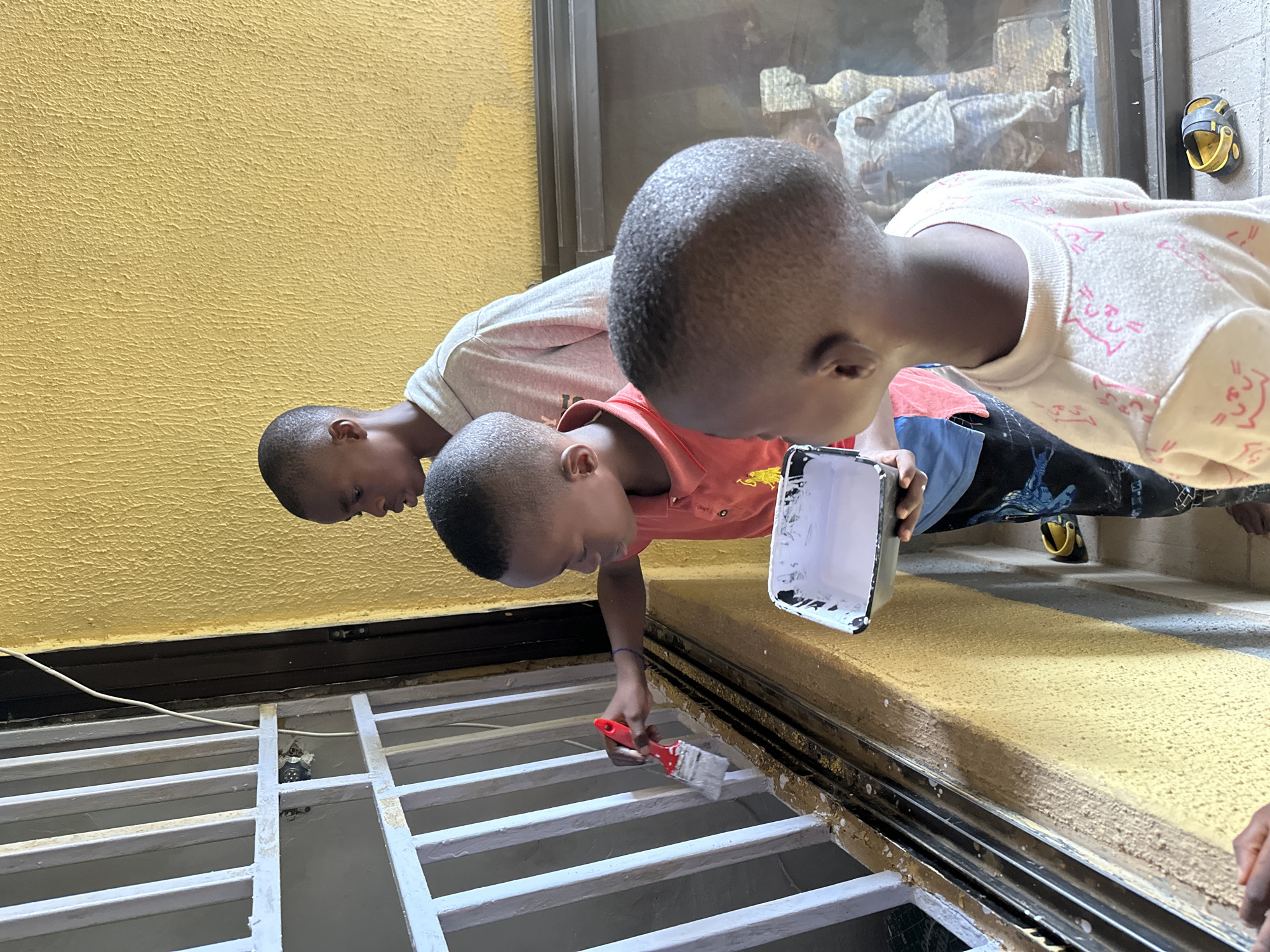 Three young boys are painting a white barred window frame in front of a yellow textured wall. The boy in the foreground on the left, wearing a red polo shirt, holds a paintbrush in his right hand and dips it into a white paint tray held in his left hand. He is focused on painting the bars. To his right, another boy, partially obscured, wears a light grey hooded sweatshirt and stands behind him, looking in the same direction. In the immediate foreground, a third boy, facing away from the camera, wears a light-colored sweatshirt with pink cat-like graphics. To the right of the boys, a framed artwork or picture is visible, showing figures in patterned clothing. The ground is uneven, with some yellow paint splatters and a pair of yellow and black shoes. The scene appears to be an outdoor or semi-outdoor space, possibly a residential area in Narayi, Nigeria.