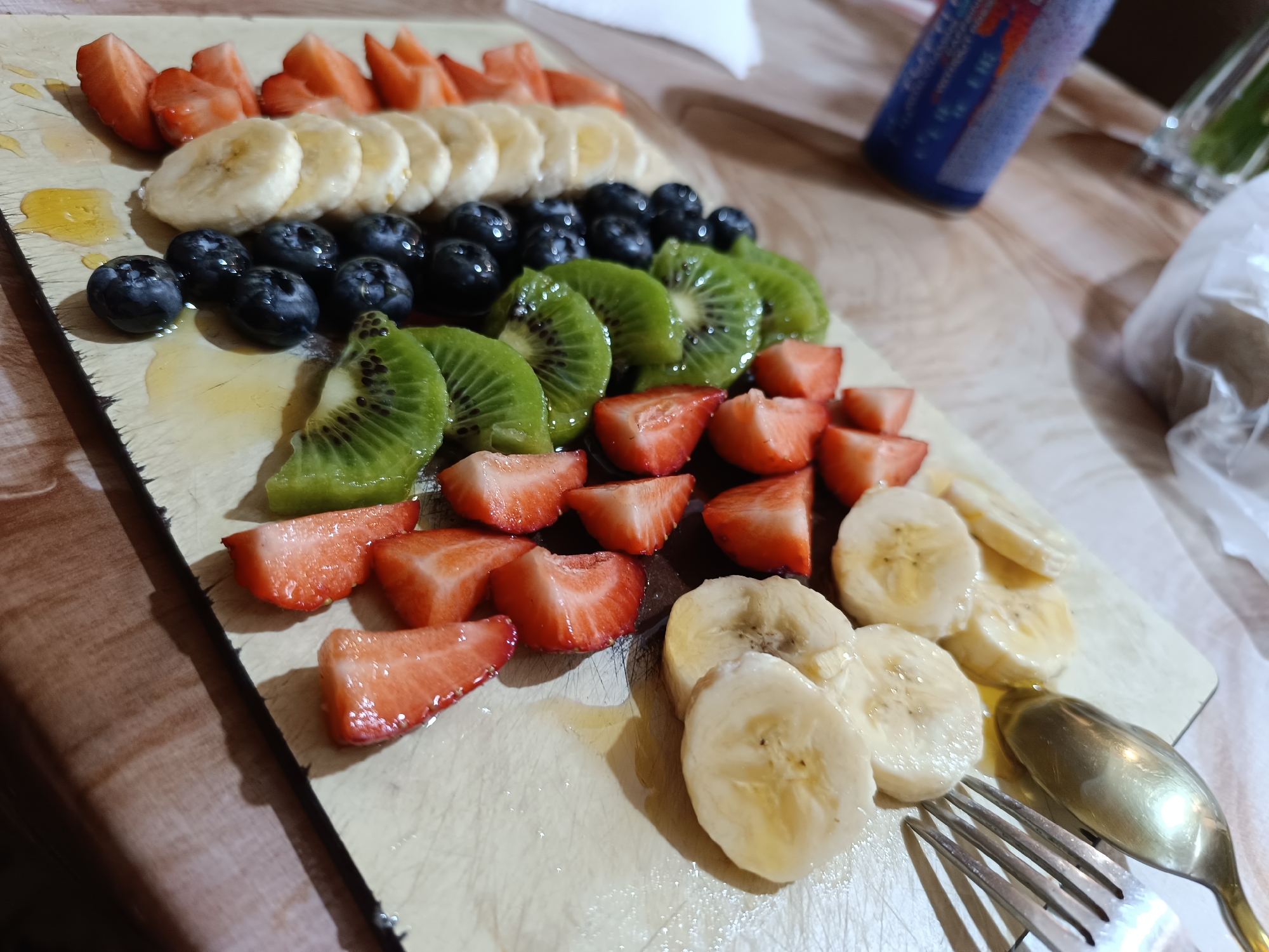 The image shows a wooden board with a variety of sliced fruits, including strawberries, bananas, blueberries, and kiwi, arranged in rows. A small pool of a translucent liquid, possibly honey, is visible on the board among the fruits. A fork and spoon rest near the bottom right corner of the board. In the background, partially blurred, are a blue can and what appears to be a glass or container with a green liquid. The scene is indoors, on a table or countertop. There is no information in the image connecting it to Pleven, Bulgaria.