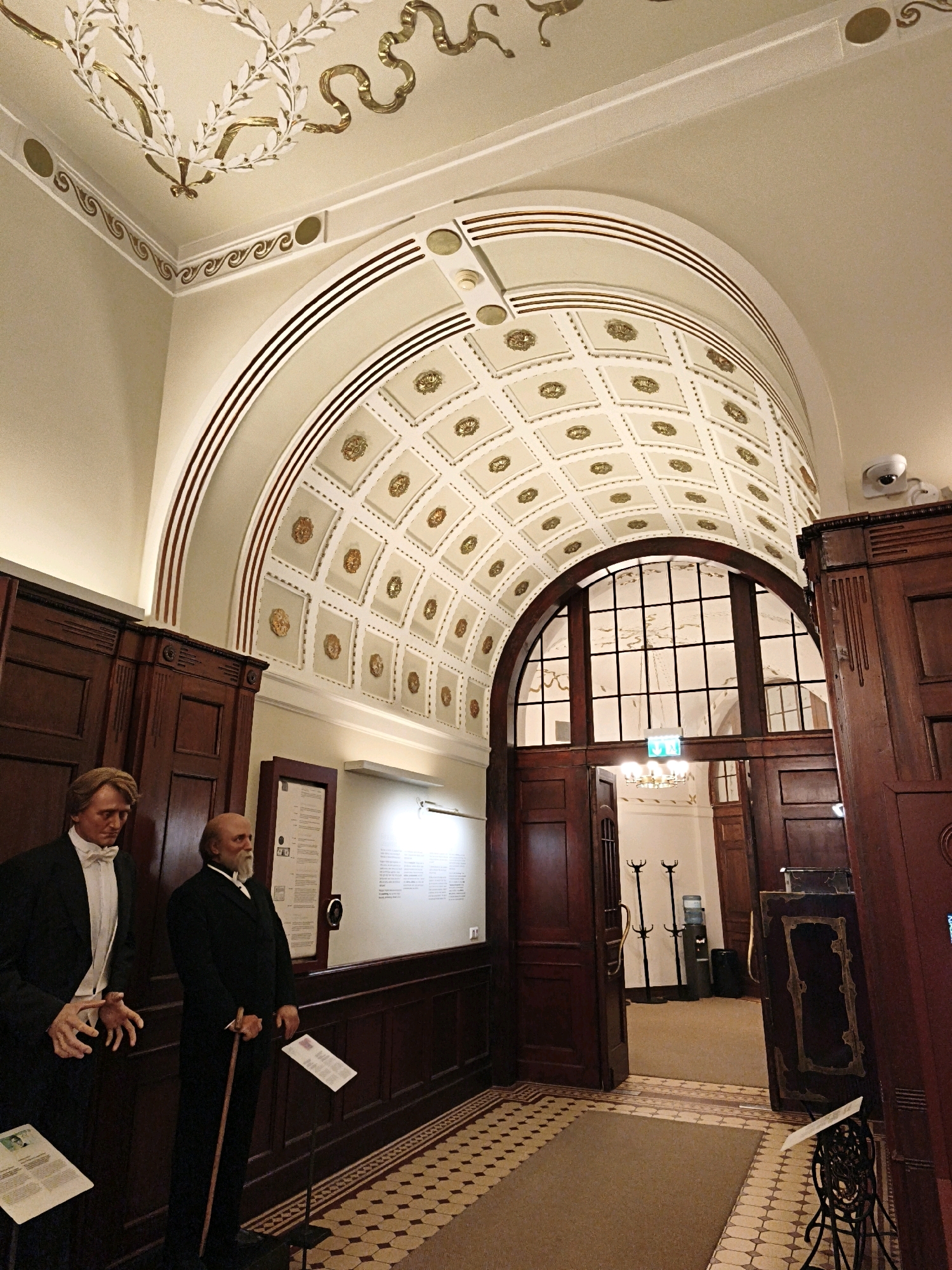 The image shows the interior of the Kiek in de Kök Museum in Tallinn, Estonia. The scene captures a grand hall with a vaulted ceiling adorned with intricate gold decorations and coffers. In the foreground, two mannequins dressed in historical attire stand on a tiled floor. The mannequin on the left wears a formal suit with a white bow tie, while the mannequin on the right is dressed in a dark suit and holds a cane. Behind them, dark wooden paneling lines the walls, and a large arched doorway with stained glass is visible in the background. The doorway leads to another section of the museum, where a chandelier and coat racks are discernible. The overall ambiance suggests a historical exhibition space.