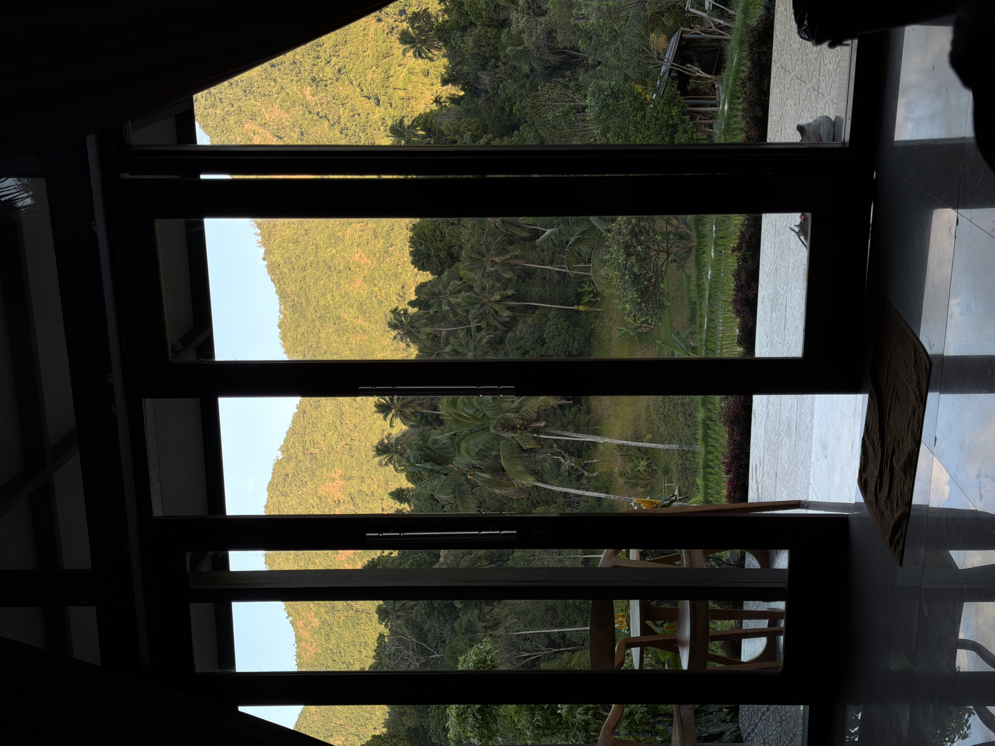 The image shows a view from inside a building looking out through large glass doors. The doors are open, revealing a lush green landscape with rolling hills covered in trees and vegetation. In the foreground, a tiled floor reflects the light from the outdoors, and a small mat is placed on it. Two wooden chairs and a table are visible on a patio area just outside the doors. Beyond the patio, there are rice paddies and more dense foliage, leading up to the hills. The sun appears to be setting or rising, casting a warm glow on the hills. A small wooden structure, possibly a shed or hut, is visible in the distance. The scene suggests a serene and tropical environment, likely in Sawan, Indonesia, given the characteristic terraced rice paddies and the tropical vegetation. There are no people clearly visible in the image.