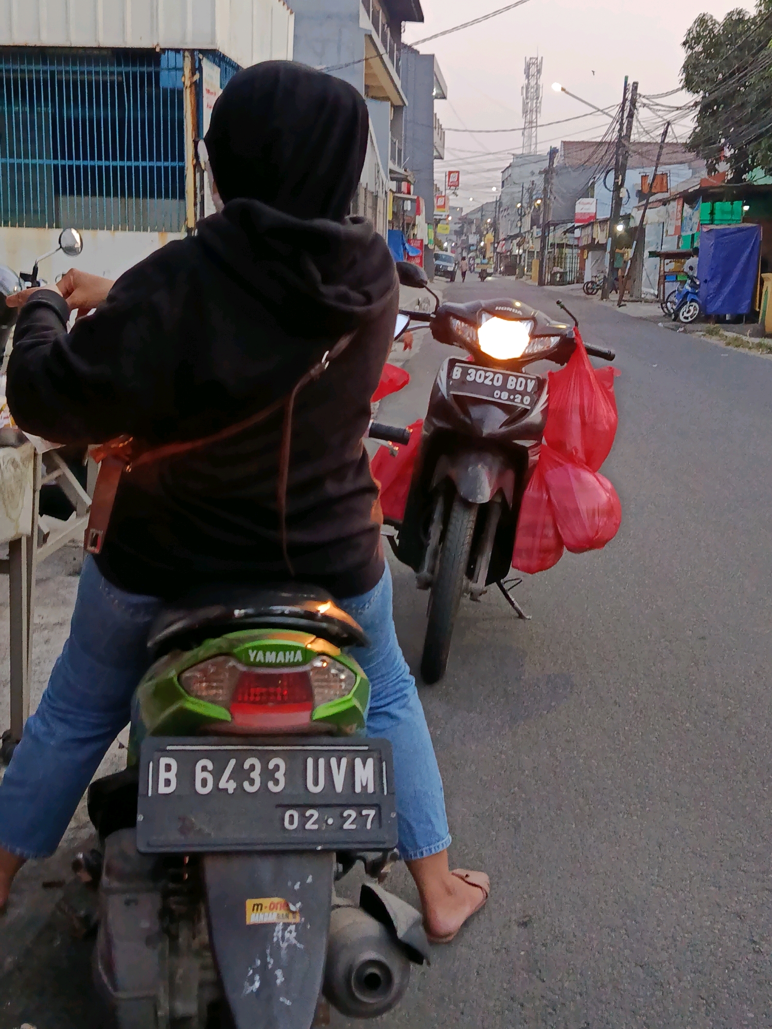 A rear view of a person wearing a black hijab and black hoodie sits astride a motorcycle in Bekasi, Indonesia. The person wears blue jeans and light brown sandals. The motorcycle has a green rear fender and a license plate reading "B 6433 UVM 02.27". Two red plastic bags hang from the handlebars of another motorcycle parked in front of the person's. This motorcycle has a license plate reading "B 3020 BDV 08.20" and its headlight is illuminated. The scene appears to be a street with buildings lining both sides. The sky is overcast and a cell tower is visible in the distance.