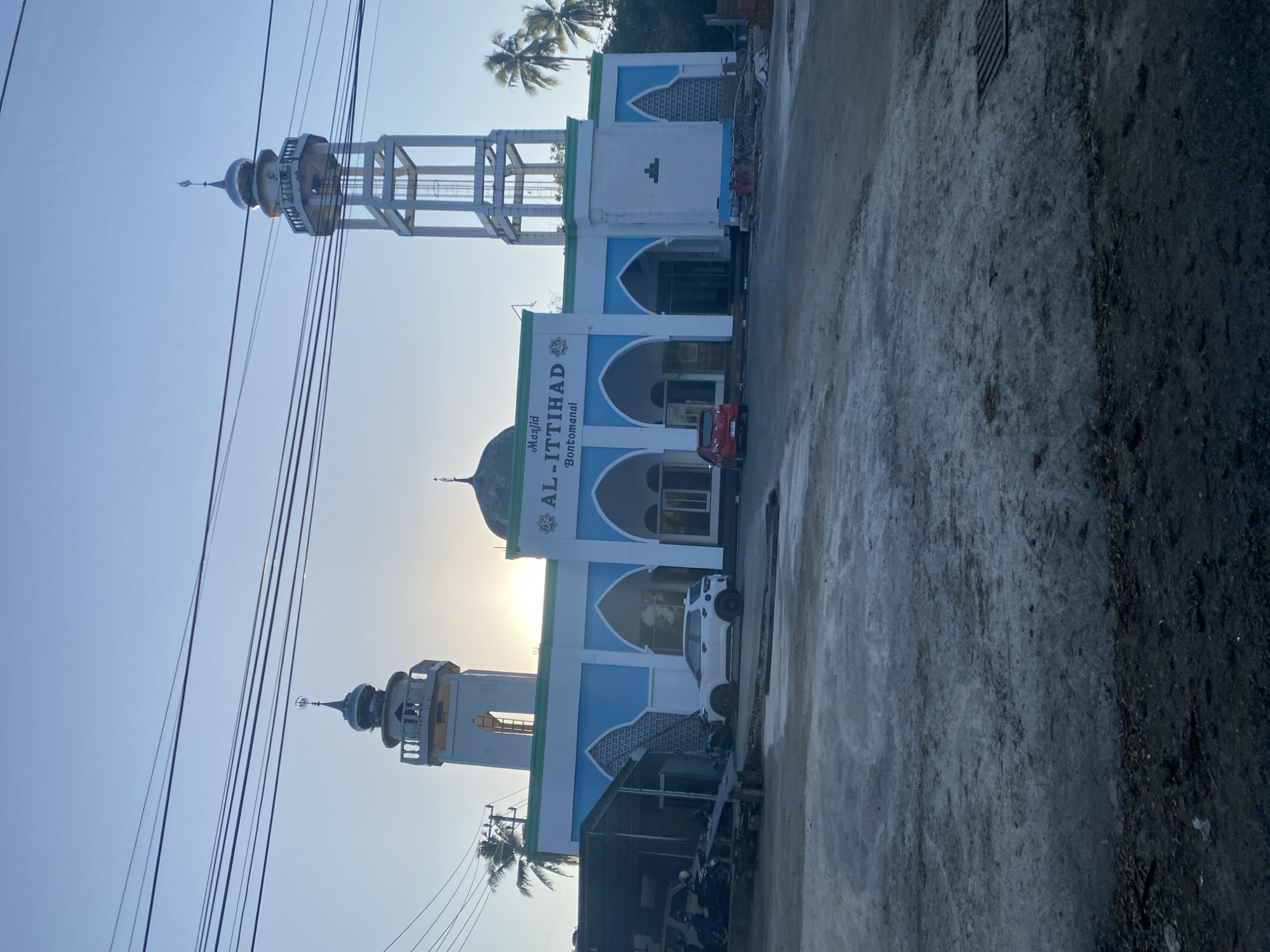 The image depicts the AL-ITTIHAD mosque in Batukaropa, Indonesia, under a clear, pale blue sky at dusk or dawn. The mosque has a white and light blue facade with arched doorways and windows. Two minarets stand tall; one on the left is shorter and topped with a dome, while the one on the right is taller and appears to be a metal frame structure with a dome at the very top. The setting sun casts a warm glow behind the left minaret. In front of the mosque, there is a paved area with a white SUV parked to the left of the entrance and a small red car parked to the right. To the far left, there's a covered area with motorcycles and some construction materials. The foreground is a rough, greyish paved surface with some debris and a drainage grate visible on the right side of the image. Palm trees are visible to the right and left of the mosque, indicating a tropical environment. Electrical wires are strung across the upper portion of the image.