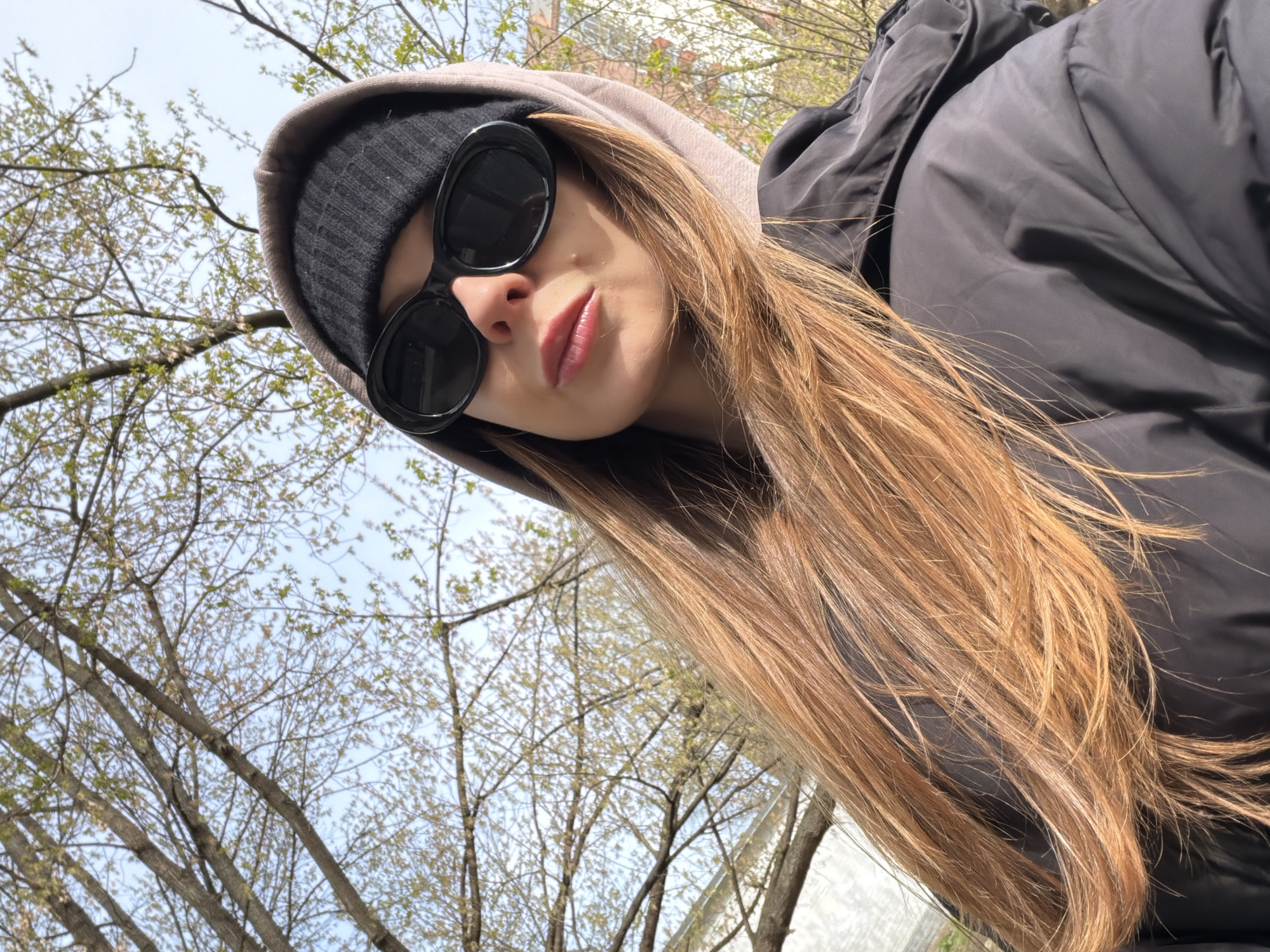 A young person is captured in a close-up, low-angle self-portrait, looking downwards towards the camera. The subject's face is partially obscured by dark, oval-shaped sunglasses and a black knitted beanie pulled down low. They are wearing a light-colored hooded sweatshirt beneath a dark, possibly black or navy, puffy winter jacket. Their long, light brown hair, appearing somewhat wind-blown or styled, cascades down the right side of the frame and over the jacket. The lips are visible beneath the sunglasses, forming a slight pout with a hint of natural or lightly applied pink lip color.
The background consists of a bright, light blue sky with numerous bare tree branches beginning to show nascent green buds and leaves, indicating a spring season. The sunlight appears strong, casting some shadows and highlighting the hair. No specific landmarks or buildings are clearly visible, but the presence of trees suggests an outdoor setting, possibly a park, street, or garden in Khimki, Russia. The overall impression is a casual, outdoor snapshot taken on a clear day. There is no visible text in the image.