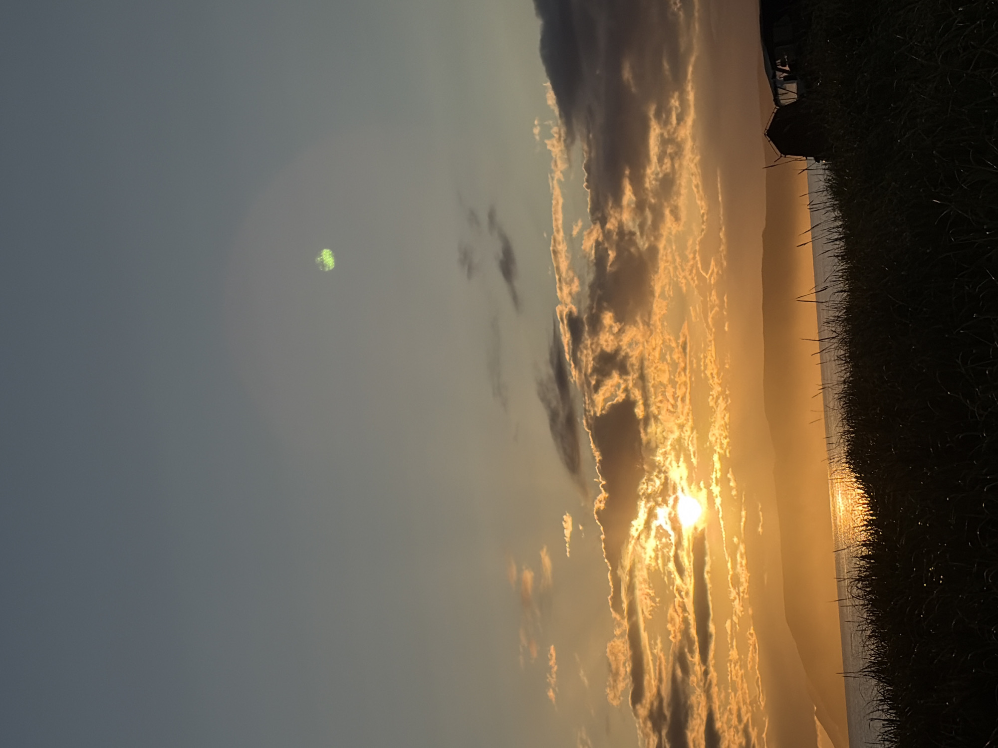 This image depicts a sunset over a body of water, likely a sea or large lake, with land formations visible in the distance. The sky is a gradient of blues and oranges, with scattered clouds illuminated by the setting sun. In the foreground, tall grass is silhouetted against the landscape. There are two dark, indistinct objects in the middle right of the frame that appear to be structures or vehicles. The location is identified as Ozerskoe, Russia, though specific landmarks within Ozerskoe are not discernible in this general landscape shot. There are no people or discernible actions in the image.