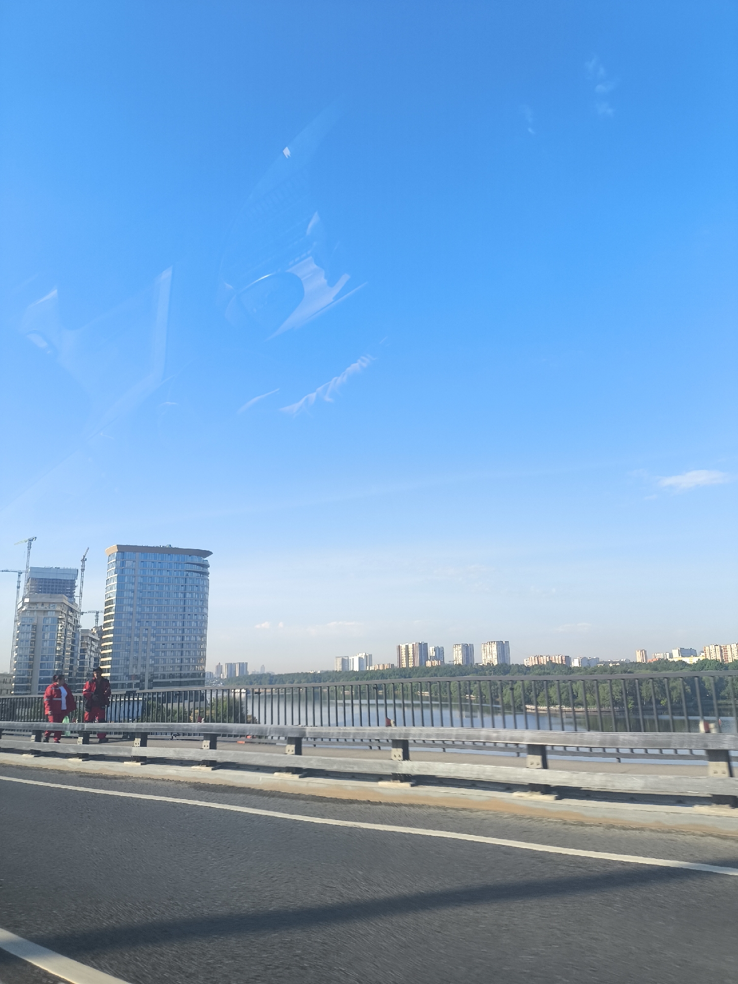 The image depicts a view from a bridge, likely in an urban setting. Two individuals wearing red work suits stand to the left of the frame, adjacent to the bridge railing. In the background, several modern high-rise buildings are visible, some under construction with cranes. A body of water, likely a river, stretches between the bridge and the buildings. Beyond the river, a distant skyline of similar buildings is discernible. The sky is clear and bright blue. The bridge itself has a metal railing and a marked asphalt road. The image is taken from a moving vehicle.