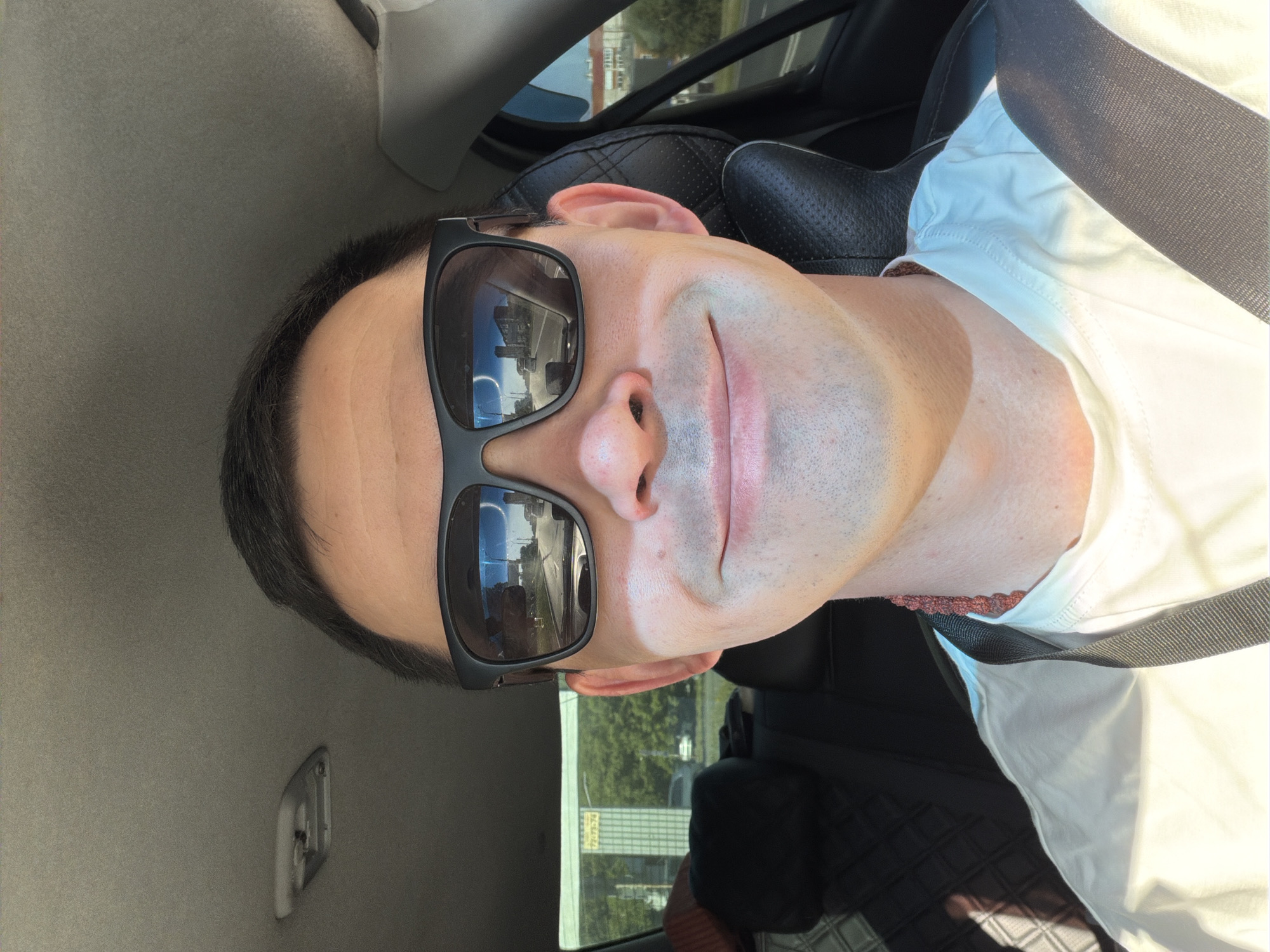 A person wearing sunglasses and a white shirt is seated in a car. The car's interior is visible, including the headrest, seat, and seatbelt. Outside the car window, a building with a sign written in Cyrillic script can be seen. The sign reads "РИЭЛТОРСКАЯ КОМПАНИЯ" at the top, and "СЕМЬЯ" at the bottom. Based on the Cyrillic writing and the presence of a building with a sign, the location is likely in a Russian-speaking area.
Given the identification of Izhevsk, Russia, the building is likely within the city limits.