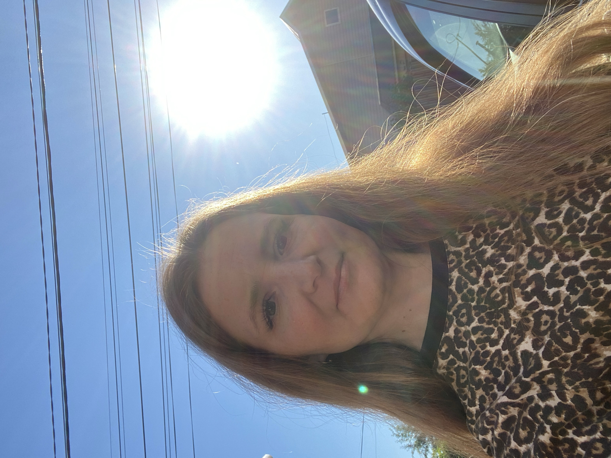 The image is a close-up, outdoor, selfie-style photograph of a young woman with long, reddish-brown hair. She is wearing a black top with a leopard print pattern. The background consists of a clear blue sky with several power lines running horizontally. The sun is bright and positioned to the upper right of the frame, causing lens flare and illuminating the woman's hair. A portion of a building and a vehicle are visible on the right side of the image. The scene appears to be in a residential or suburban area. The location in the city of Chaikovsky, Russia, is not determinable from the visual information in the image.