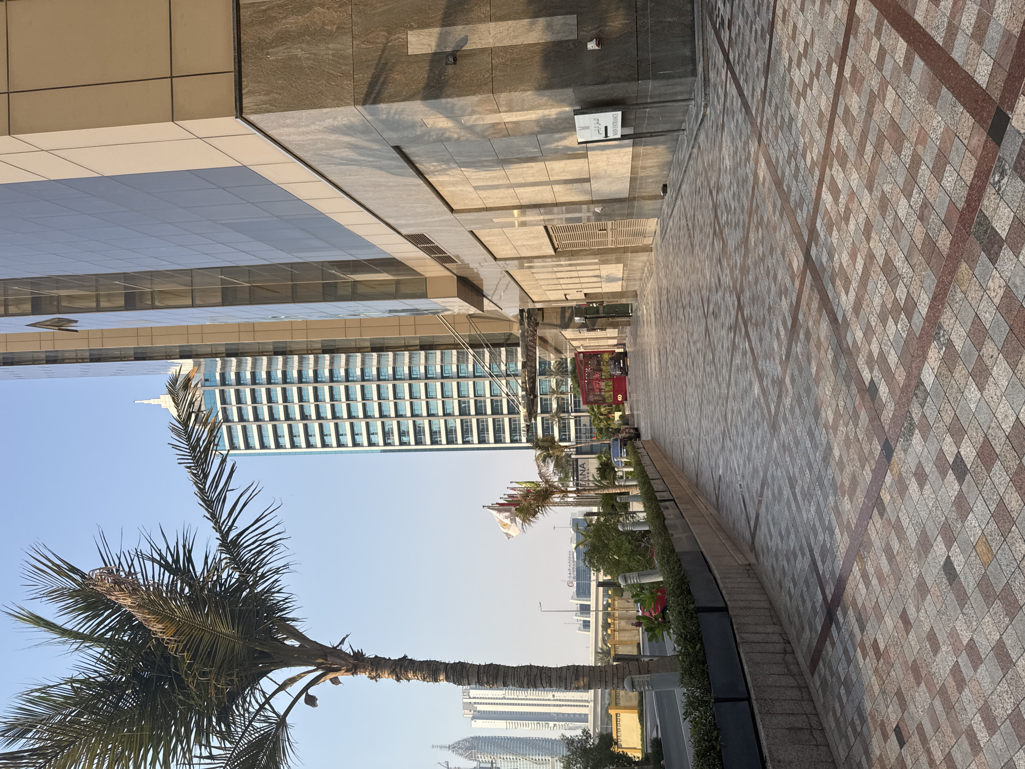 The image shows a view looking down a paved walkway in front of a modern building with a glass facade. A palm tree is on the left side of the frame, with a building in the background that appears to be under construction. Further in the background, other high-rise buildings are visible. Flags are flying on poles near the entrance of the building on the right. A red vehicle is parked on the street in the distance. The walkway is paved with patterned tiles. The scene appears to be in a city setting, likely an urban area in Dubai, United Arab Emirates, given the architectural style and presence of palm trees. The lighting suggests it is daytime, possibly late afternoon given the shadows. There are no people actively engaged in any interactions or notable actions visible in the frame, though there may be figures in the distance.