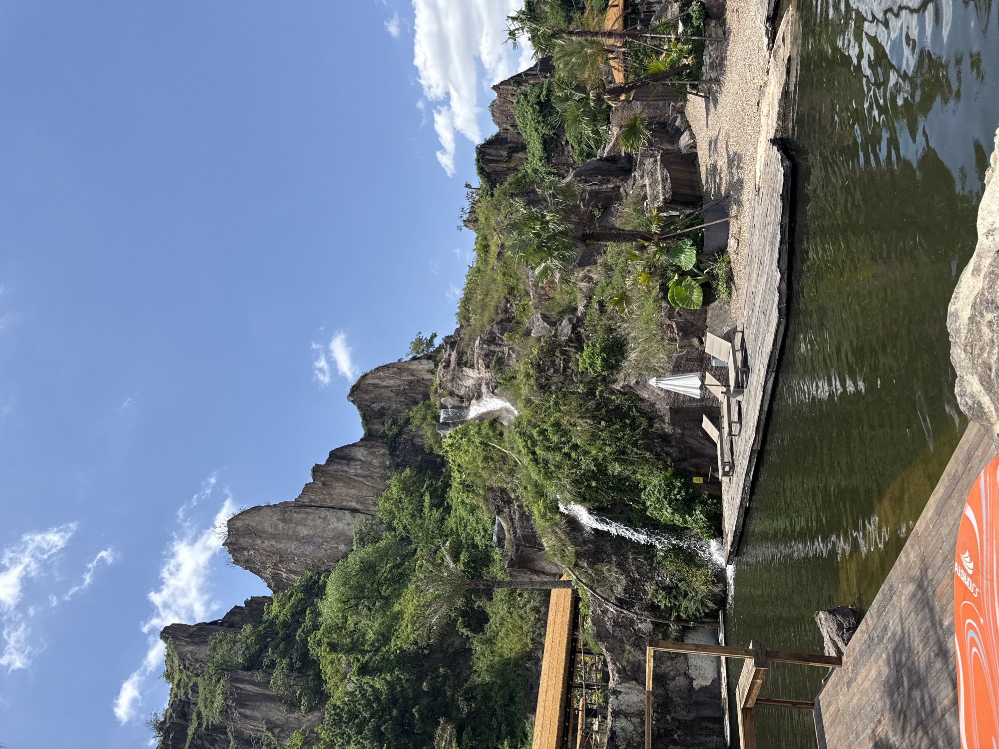 The image captures a man-made landscape with a rocky, heavily vegetated hill featuring a waterfall flowing into a constructed pond or lake, likely within a recreational or resort setting.
**Environment:** The foreground contains a wooden deck adjacent to a body of water. The water's surface shows some ripples, suggesting a slight breeze or movement. Behind the deck, a steep hill or artificial rock structure rises, covered in dense greenery consisting of various trees, shrubs, and other foliage. Two waterfalls cascade down the hill into the pond. The sky is blue with scattered white clouds.
**Objects:** A white parasol and two lounge chairs are placed on the wooden deck. An orange and white object with text visible is in the right foreground. Some construction or support structures of wood and metal can be seen along the base of the hill.
**Action:** Water is flowing from the top of the hill in the form of a waterfall and emptying into the pond.
Given the reference to the Keqiao District, China, the specific location could potentially be a tourist resort or a landscaped area designed to resemble a natural environment.