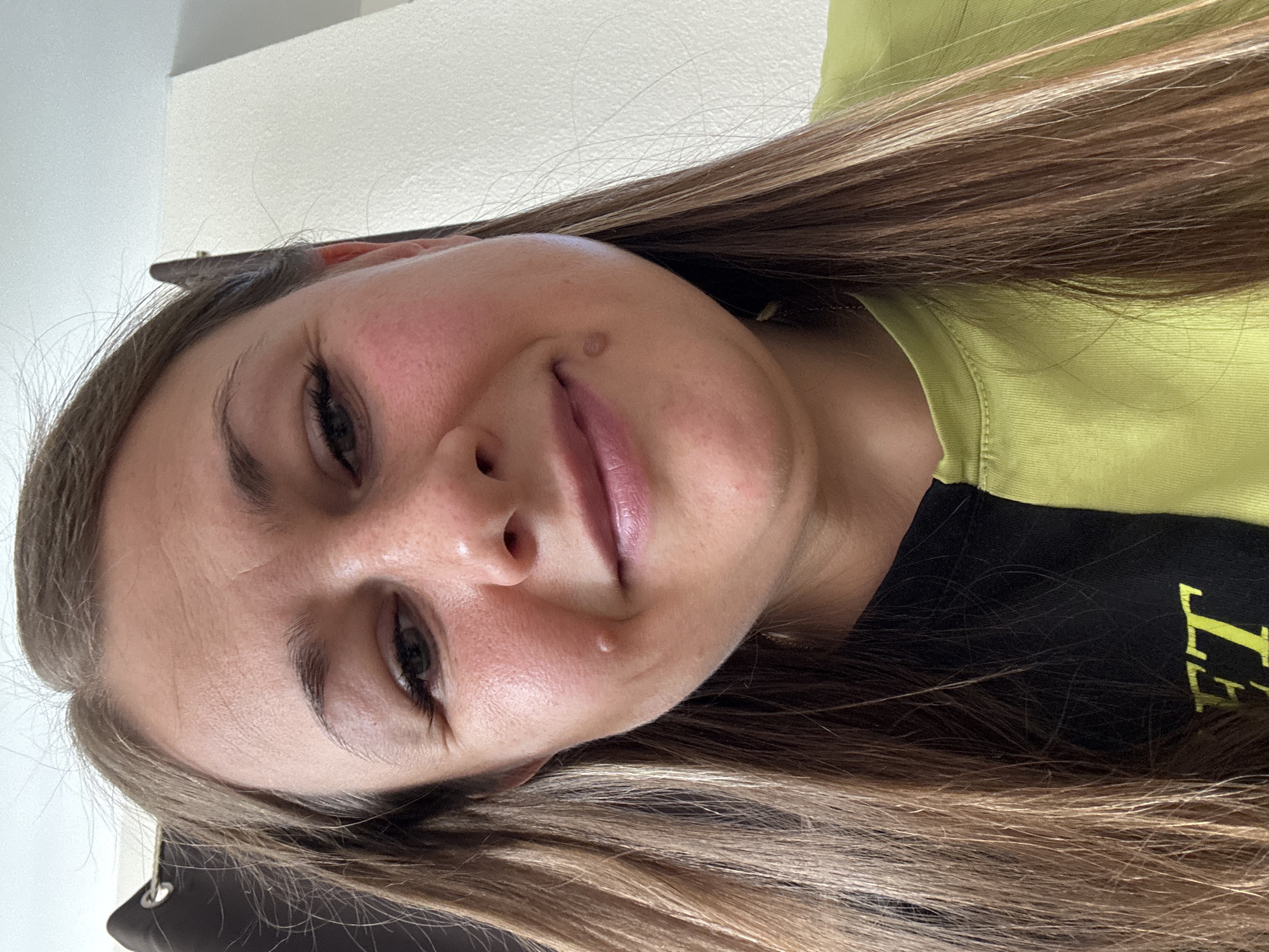 A close-up, low-angle, selfie-style shot captures a Caucasian woman's face as she looks down and slightly to her left. Her face is angled towards the camera, with her head tilted down. She has long, straight, brown hair with blonde highlights, which frames her face and cascades down her shoulders. She is wearing a split-colored t-shirt, with the left side black and the right side a bright lime green. The background consists of a light-colored wall and a dark curtain. The woman's facial features are prominent, including her eyes, nose, and mouth, as well as two moles on her right cheek. Her expression is soft, with her eyes partially closed and her mouth slightly open in a subtle smile. The lighting is natural and casts a slight shadow on her face, highlighting her skin texture. There is no indication of the city of Uyutnoye, Ukraine, or any specific environmental details within the image.