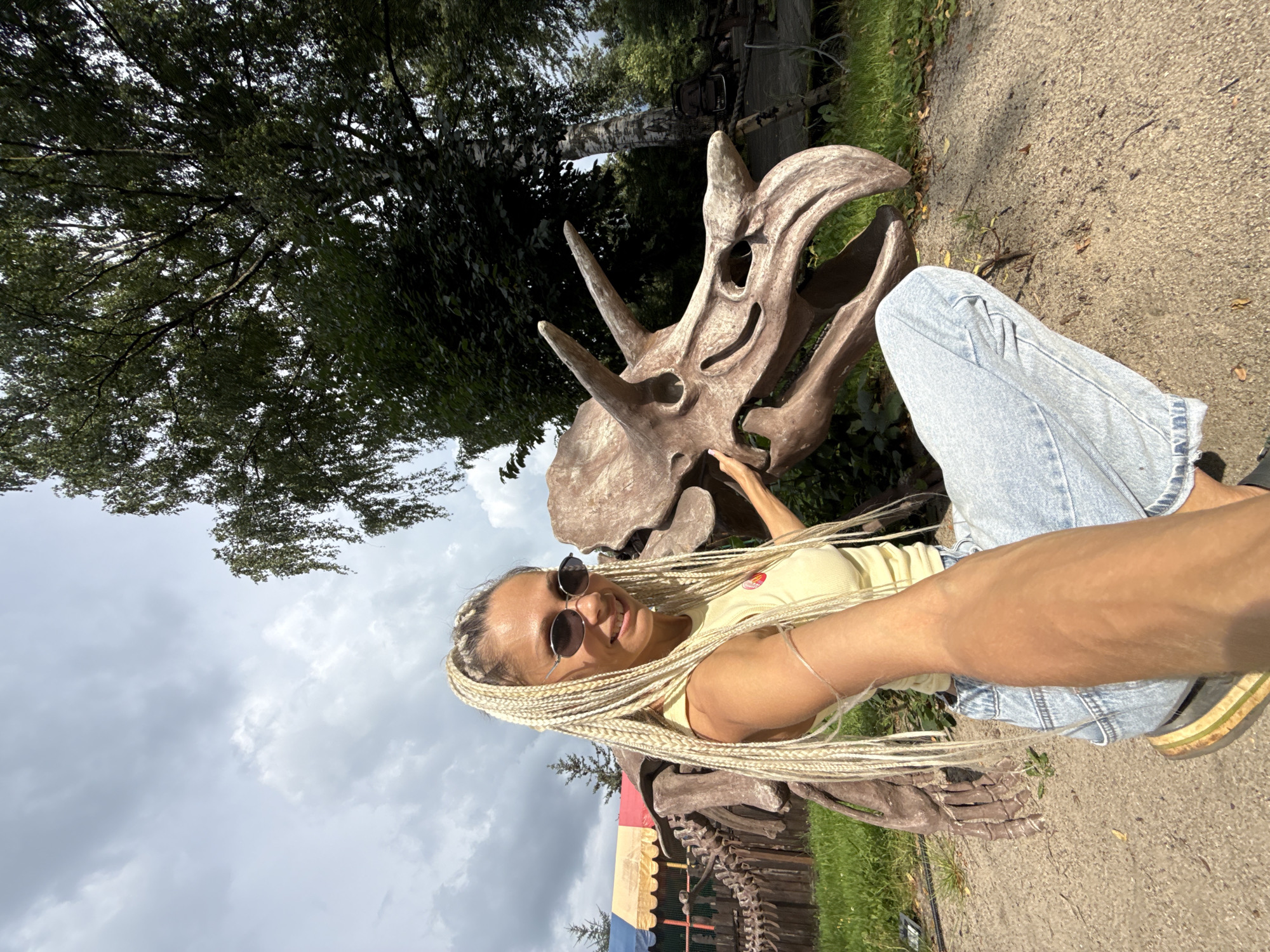 In Zhukovo, Russia, a fair-haired woman with long blonde braids wears a yellow top and blue jeans. She is crouched next to a large, brown dinosaur skeleton, her left hand resting on its skull. The skeleton is positioned outdoors on a sandy path, with green grass and trees in the background. A colorful tent is visible to the left of the scene. The sky is overcast with white and gray clouds.