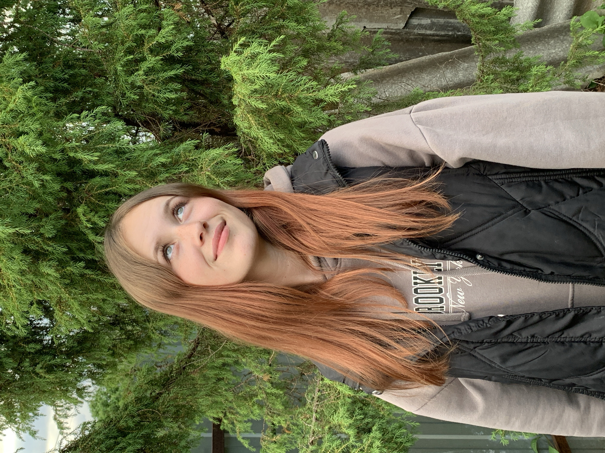 A young woman with long, reddish-brown hair stands outdoors in Фоки, Russia. She is positioned slightly off-center to the left, looking upwards and to her right with a subtle smile. Her eyes are light-colored, and she has a few visible freckles. She wears a light taupe or beige hooded sweatshirt with "BROOKLYN" and "New York" printed in dark letters on the chest, layered under an unzipped black quilted vest. The background is dominated by dense, vibrant green coniferous foliage, likely juniper, with branches extending into the frame from above and behind the subject. A metallic fence or corrugated sheet is visible on the far left, and a weathered, grey corrugated structure or wall is partially visible through the foliage on the right side of the frame.