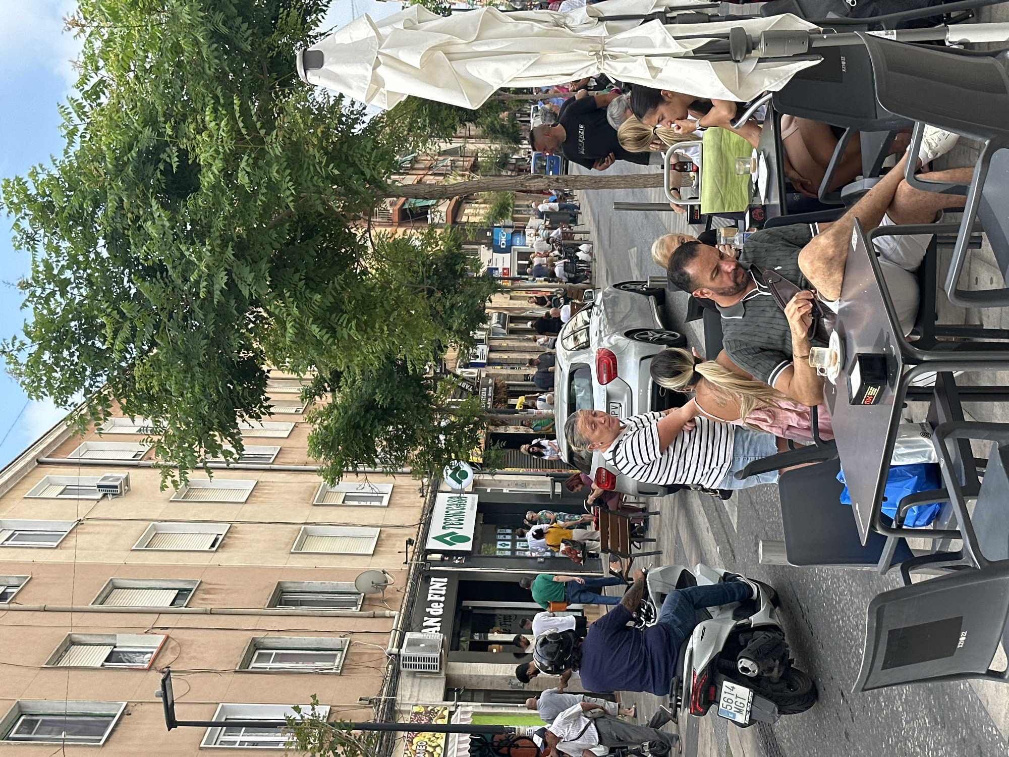 The image, tilted 90 degrees clockwise, captures a vibrant outdoor street scene, likely in a European city. Numerous people are present, primarily seated at tables under large white sun umbrellas, enjoying an outdoor cafe or restaurant. They appear engaged in casual conversation, drinking beverages, and some are looking at their phones. Other individuals are seen walking on the sidewalk or standing near the buildings.
The setting features multi-story buildings lining the street, with residential or commercial structures displaying numerous windows, some with blinds or shutters partially open. Lush green trees provide shade along the street. Street furniture includes black tables and chairs, and a white car and a silver motorcycle are visible on the road, with a person seated on the motorcycle.
It is clearly daytime, with bright sunlight creating visible shadows, suggesting fair weather and a clear sky. The general atmosphere is relaxed and social. A Spanish license plate format ("5614 MGT") on the motorcycle, along with the architectural style and cafe culture, indicates a location in Spain.
Visible text includes:
* "AN de FINI" on a storefront.
* A green logo and text "TECNOCASA" (OCR suggests "PRONOGASA").
* "DIG" on a sign.
* "MCKENZIE" on a man's black t-shirt.
* The motorcycle license plate "5614 MGT".
* "PV" partially visible on a car's license plate.
* "XZ1 22" and "XZ1022" on the backrests of chairs.