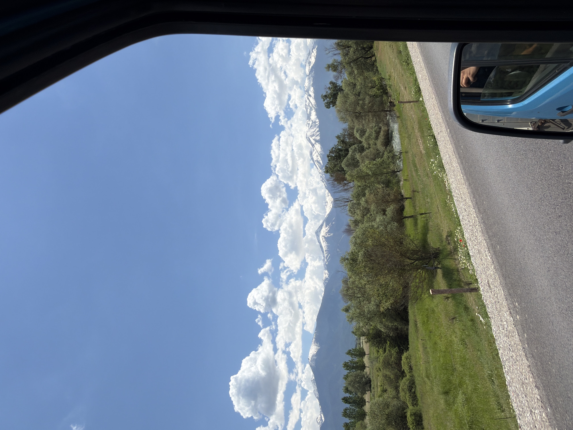 The image depicts a scene viewed from inside a vehicle, possibly a car or truck, traveling on a paved road. A side-view mirror is visible, reflecting the side of the vehicle. Outside the window, the landscape features a clear blue sky with numerous white, puffy clouds. A range of mountains with snow-capped peaks is visible in the distance. In the foreground, green fields and trees line the roadside. It is daytime, with bright sunlight illuminating the scene. The location cannot be determined based on the provided information.