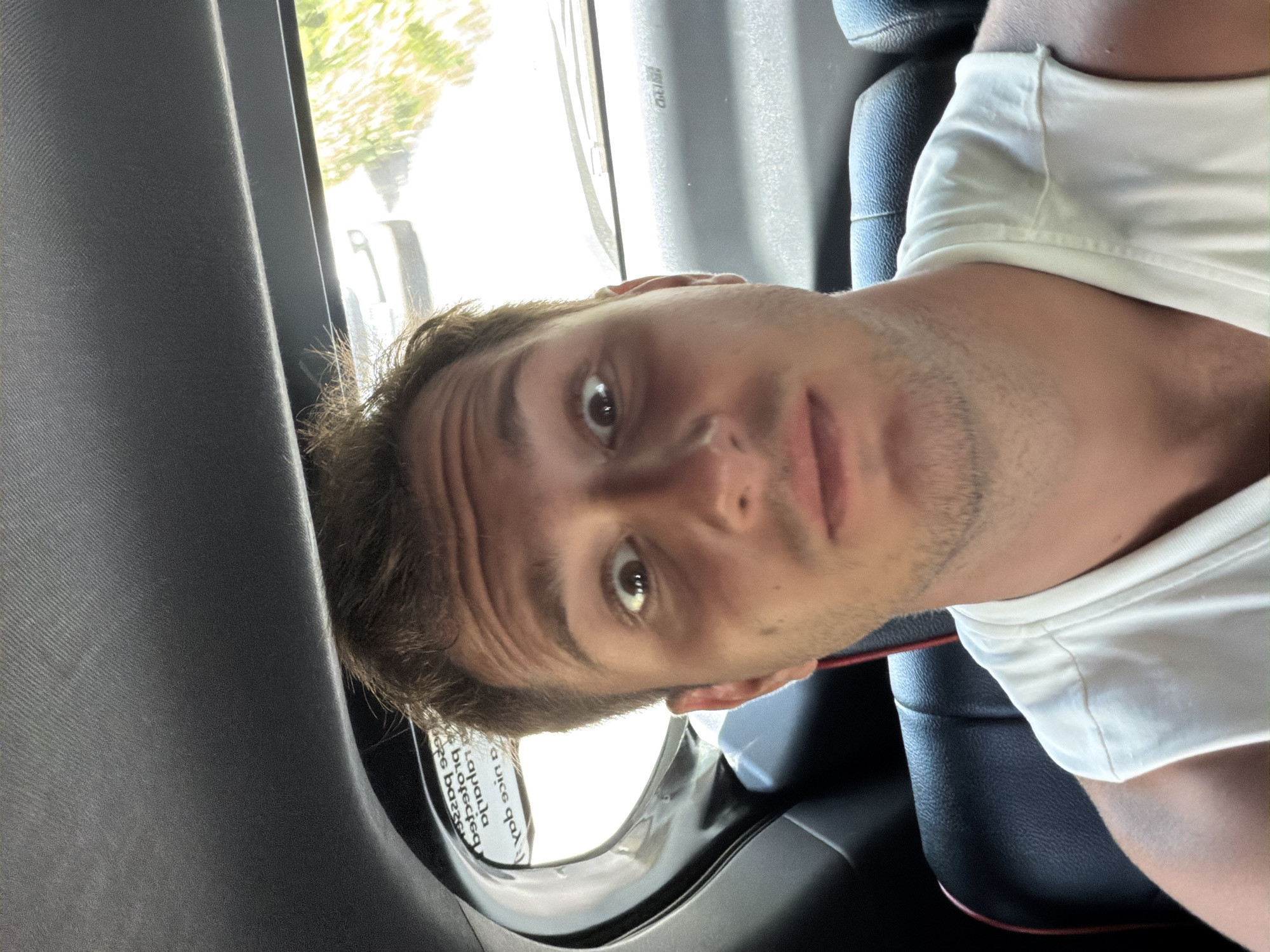 A man with brown hair and light skin is seated in the back of a car. He is wearing a white t-shirt. He is looking directly at the camera with a neutral expression. Through the car window, blurry foliage and a building can be seen. A sticker with text is visible on the car window. The scene suggests the man is a passenger in a vehicle, likely traveling in or around the city of Batununggul, Indonesia.