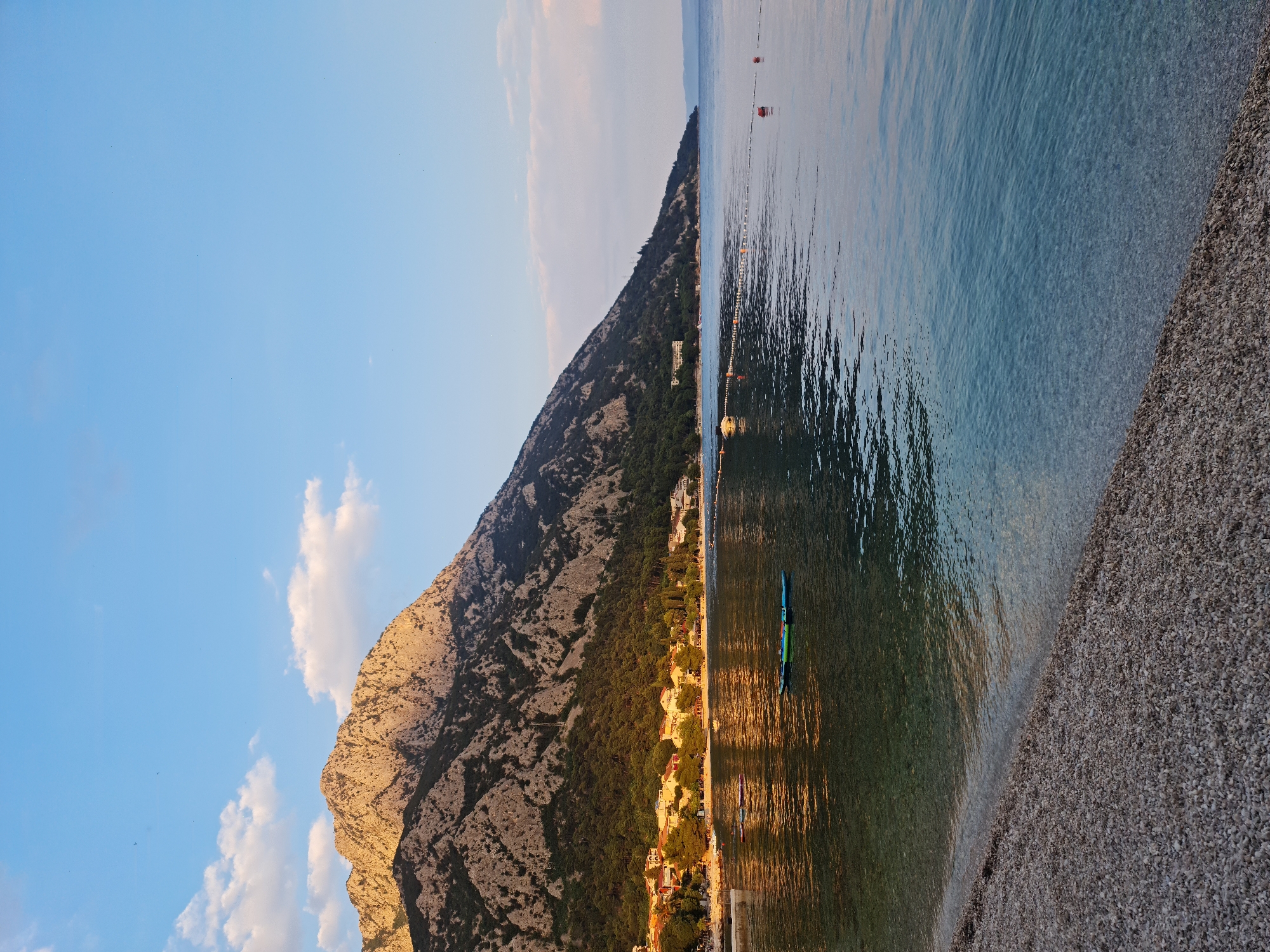 The image depicts a serene coastal scene at sunset in Općina Gradac, Croatia. A rugged, tree-covered mountain dominates the left side of the frame, its peaks illuminated by the warm golden light of the setting sun. Below the mountain, a line of white buildings with red roofs is nestled along the shoreline, suggesting a small town or village. The foreground is a pebbled beach, with the gentle waves of the clear turquoise Adriatic Sea lapping at the shore. Several buoys mark a roped-off swimming area in the water, and a few inflatable paddleboards are visible, floating calmly on the surface. The sky above is a soft blue, with scattered white clouds catching the last rays of sunlight. The overall atmosphere is one of tranquility and natural beauty.
