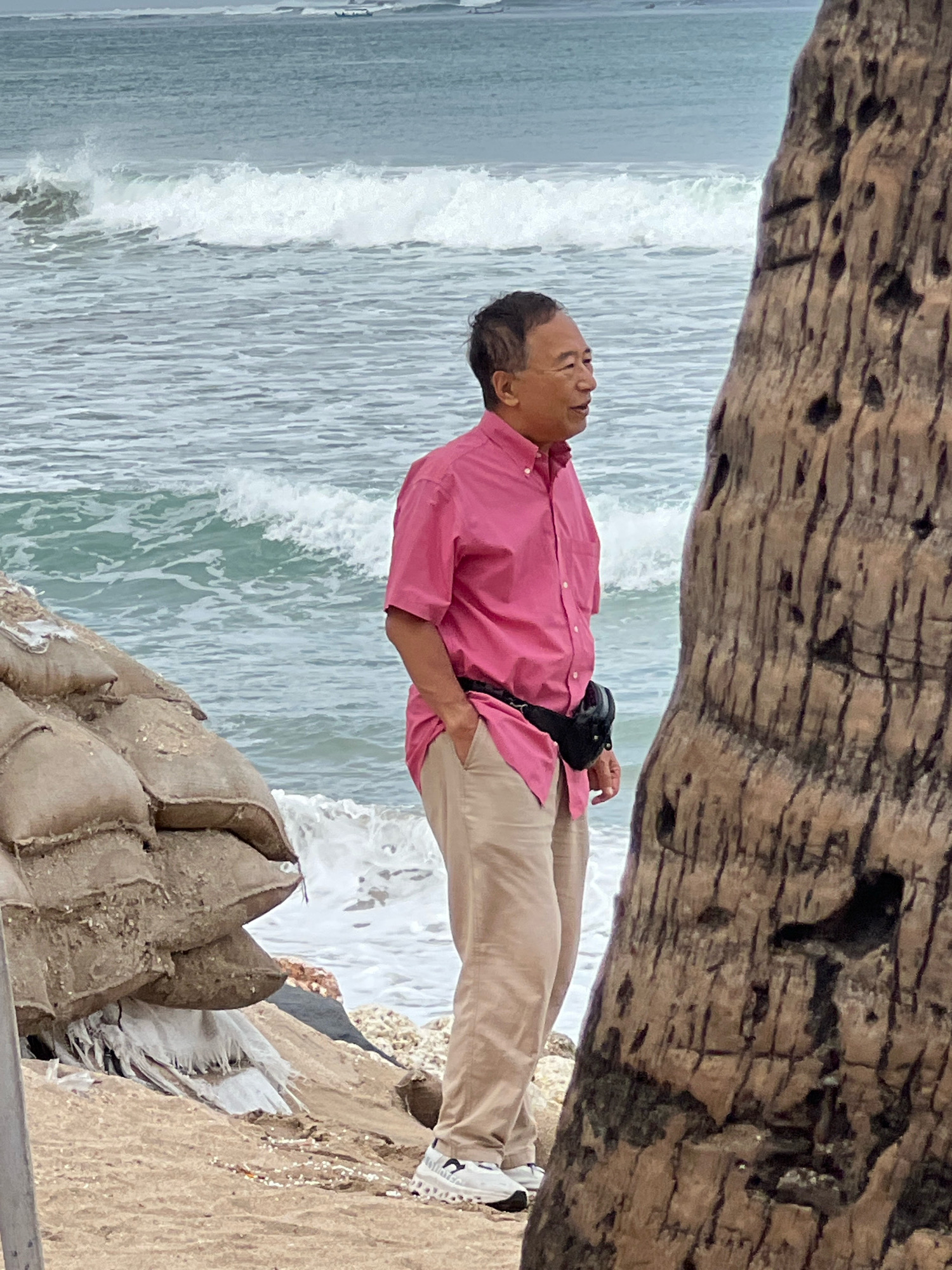 A man in a pink short-sleeved button-down shirt and tan pants stands on a beach in Kuta, Indonesia. He is looking to his right, with his left hand in his pocket. A black fanny pack is around his waist. He is wearing white sneakers. In the foreground, a portion of a large tree trunk is on the right side of the frame. To the left of the man, a pile of sandbags forms a barrier. The ocean with waves is visible in the background, and the sky is overcast.