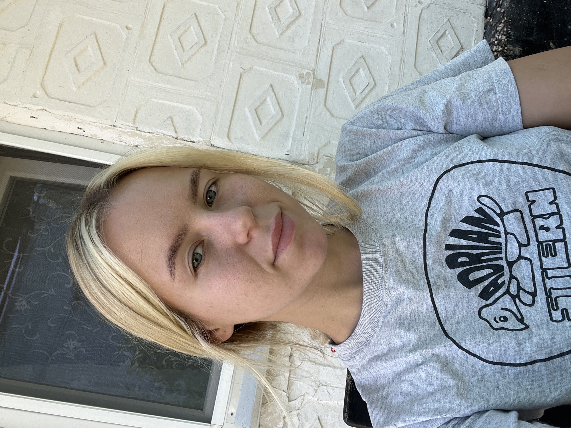 A medium shot of a blonde woman sitting outdoors, looking at the camera. She is wearing a gray t-shirt with a black oval graphic that contains the text "ADRIAN STERN" and a drawing of a turtle. The woman has light skin and light green eyes. Her hair is shoulder-length and parted to the side. She is sitting in front of a white, textured wall and a window. The lighting suggests it is daytime.