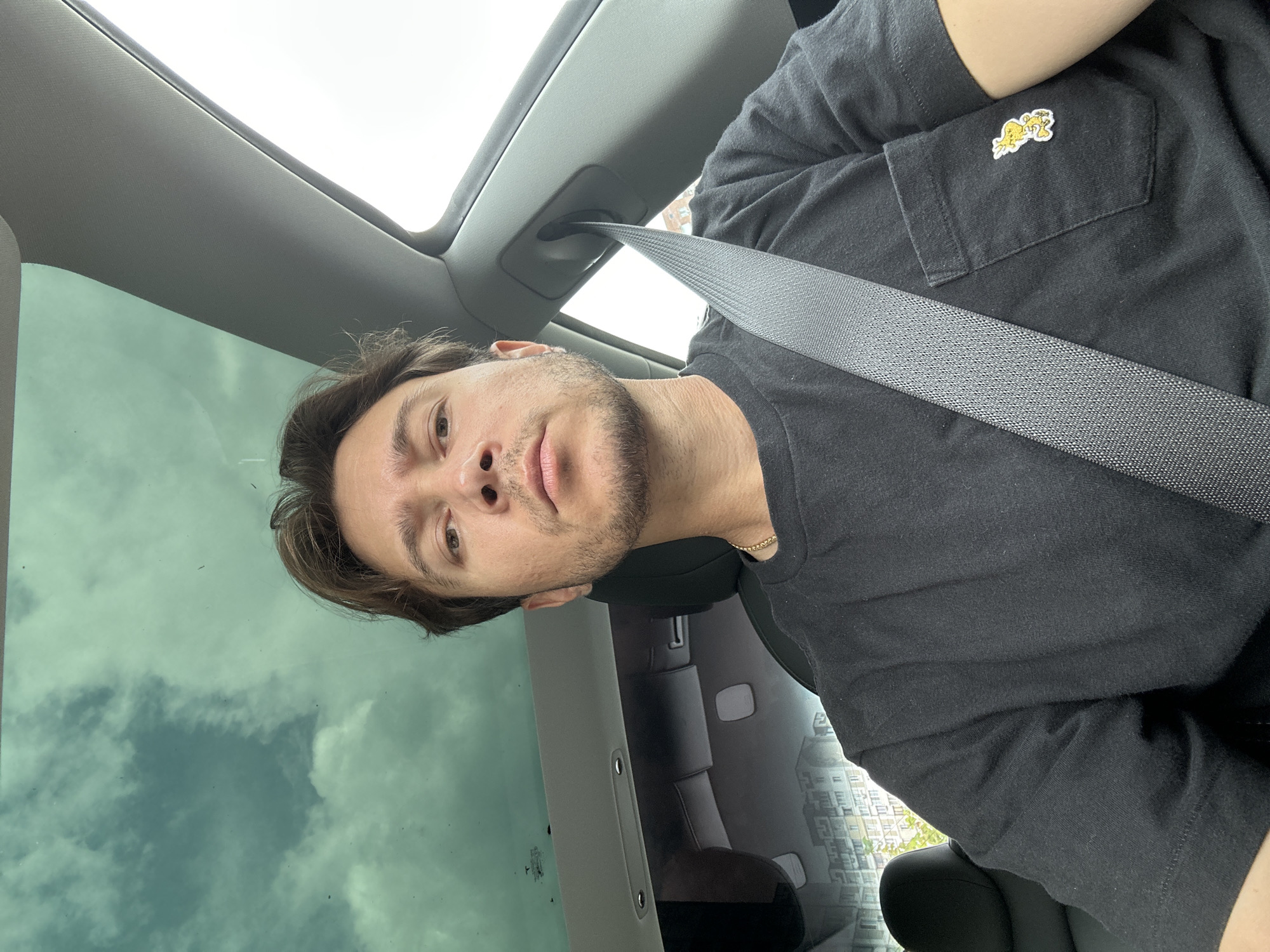 A man with short, brown hair and stubble is sitting in the driver's seat of a car. He is wearing a black t-shirt with a small yellow cartoon character patch on the breast pocket and a gold chain. The car has a sunroof that shows a cloudy sky, and through the rear window, a multi-story apartment building is visible, suggesting an urban setting in Kyiv, Ukraine. The man is looking directly at the camera, and his seatbelt is fastened across his chest. The overall scene depicts a person in a vehicle, likely in an urban environment.