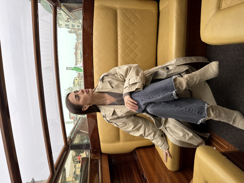 A single female subject is seated inside a water taxi traversing a canal in Venice, Italy. She occupies the left side of a yellow diamond-quilted leatherette bench seat, positioned in a three-quarters view facing right and slightly upwards. The subject has long dark brown hair and wears a light beige trench coat over a brown V-neck sweater, dark grey wide-leg jeans with frayed hems, and light beige suede mid-calf boots. Her legs are crossed, with the right over the left, and her right hand rests on her left knee. Her expression is neutral, her gaze directed towards the distant cityscape.
The interior of the water taxi features polished wooden paneling, a dark grey floor mat, and a curved transparent glass roof allowing views of the sky and surroundings. Outside the boat, under an overcast sky, a panoramic view of historic Venetian architecture is visible, including multi-story buildings, a prominent campanile, domes, and a white flagpole bearing the Italian flag (green, white, red). The scene depicts transit through a characteristic Venetian waterway.