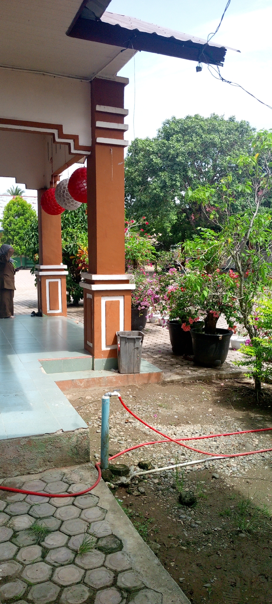 The image depicts an outdoor scene in Baratpayaitek, Indonesia, likely in front of a building. A person wearing a brown uniform and a headscarf stands on a porch, looking upwards. The porch area features hexagonal paving stones and is supported by columns. Two decorative spherical ornaments, one red and white and another red, are hanging from the porch ceiling.
Beyond the porch, a garden area is visible with several potted plants, including flowering bougainvillea in pink and white. A large tree with green foliage dominates the background. A gray trash can is placed near one of the porch columns, and a blue pipe with a red hose attached is situated on the ground in the foreground, connected to what appears to be an outdoor faucet. The ground is covered with gravel and some sparse vegetation. The sky is bright and clear, suggesting daytime.