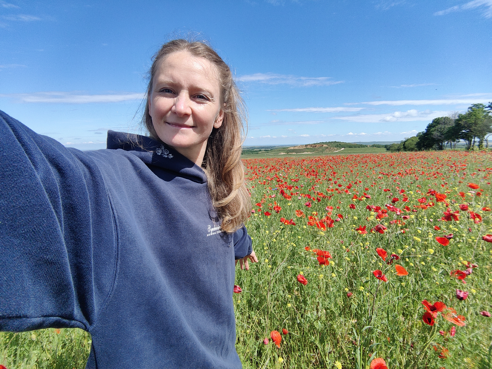 A woman with shoulder-length blonde hair takes a selfie in a field of red poppies under a clear, blue sky with scattered clouds. She wears a dark blue hooded sweatshirt. The location is Крымскорозовское сельское поселение, Ukraine, indicated by the agricultural landscape. In the background are rolling hills and trees.