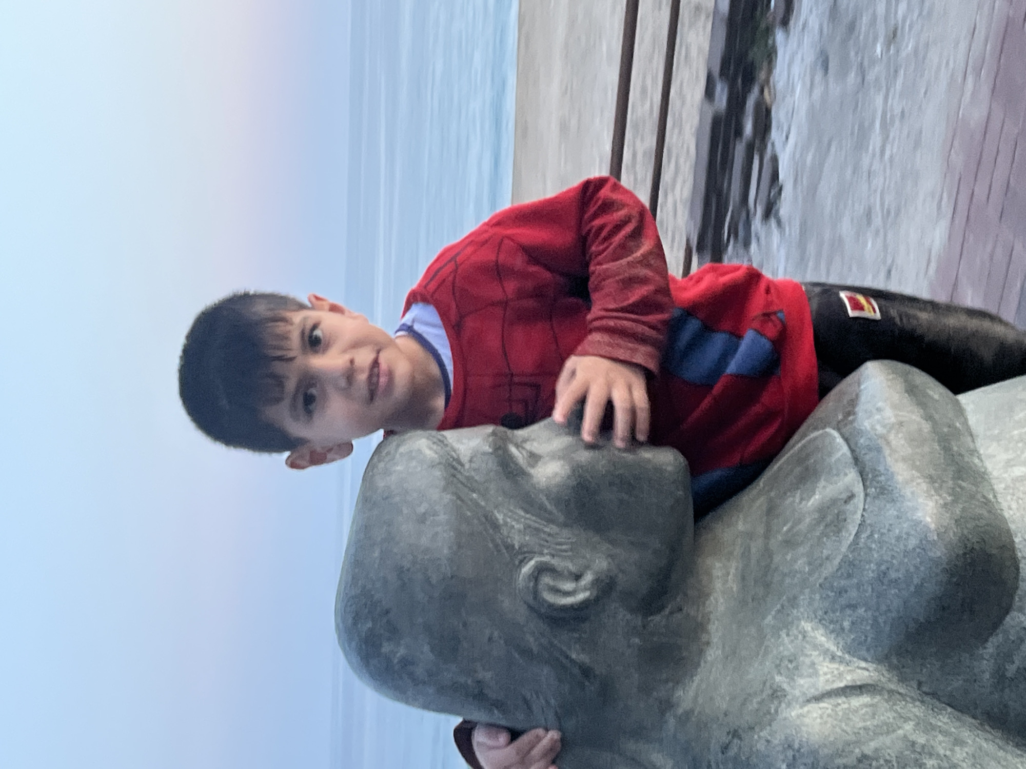 The image depicts an outdoor scene in Alanya, Turkey, featuring a young boy interacting with a large, stylized sculpture. The original image is rotated 90 degrees counter-clockwise.
A young boy, likely of elementary school age, is the central human subject. He has dark hair and a gentle smile, looking directly at the viewer. He is wearing a red, long-sleeved shirt with blue sections and black web-like patterns, suggestive of a Spiderman costume. His hands are resting on the head and upper body of the sculpture, indicating playful engagement.
The sculpture is a large, smooth, grey form resembling stone or concrete, depicting a stylized human head and upper torso in a reclining position. Its features are abstract and serene.
In the background, a wide expanse of calm water, likely the sea, stretches out with a light blue-grey hue, suggesting either an overcast day or the soft light of dawn or dusk. To the right of the boy and sculpture, a darker, textured area, possibly a pier or walkway made of wood or stone, is visible, with some vertical structures that could be railings. The overall atmosphere is peaceful and engaging. There is no discernible text visible in the image.