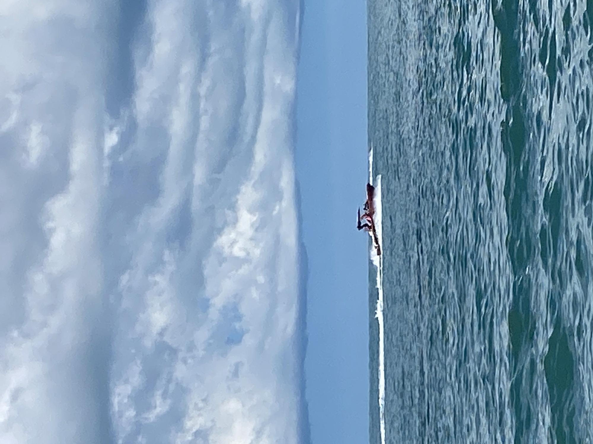 The image shows a jet ski with two people on board, moving at speed across the choppy blue water of the sea. The jet ski is dark red in color. In the background, the sky is filled with dramatic, dark grey clouds, suggesting an impending storm. The scene is set against a vast expanse of sea and sky, with no visible land or structures, making it difficult to pinpoint a specific location within Sochi, Russia, without further context. The water is a deep blue-green, with white spray trailing behind the jet ski, indicating its rapid movement. The clouds are heavy and textured, with lighter patches suggesting some diffused sunlight breaking through. The overall impression is one of dynamic motion on the water under a turbulent sky.