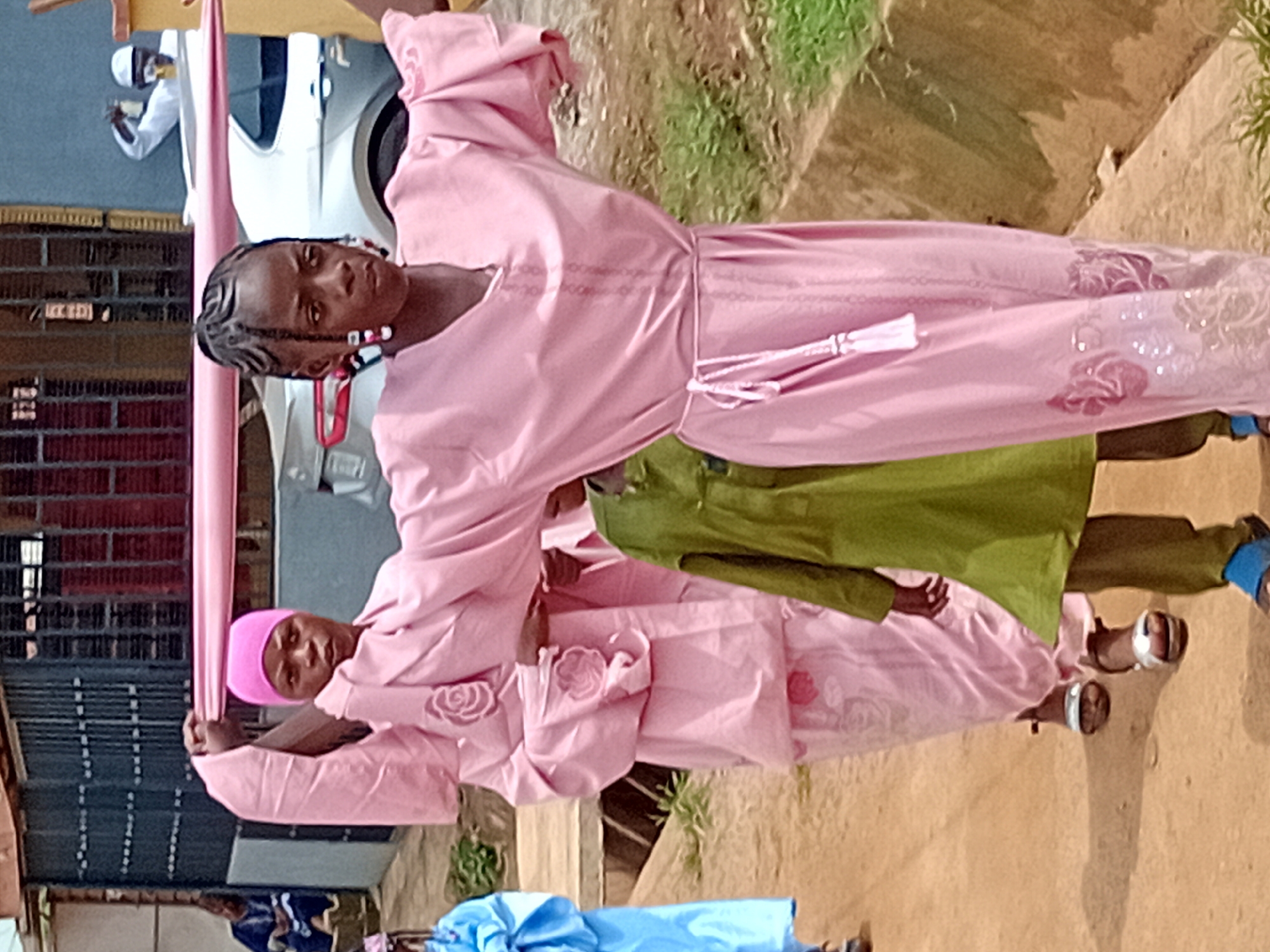 In Ikorodu, Nigeria, the image shows several people outdoors, likely on a street. A young woman with braided hair, wearing a light pink robe, stands in the foreground. Another woman in a similar pink robe and a pink head covering is beside her. An individual in a green garment is also present. They appear to be associated with an event, possibly religious. A white vehicle is partially visible in the background, along with a fence. The ground is dirt or sand, and there is some vegetation nearby.