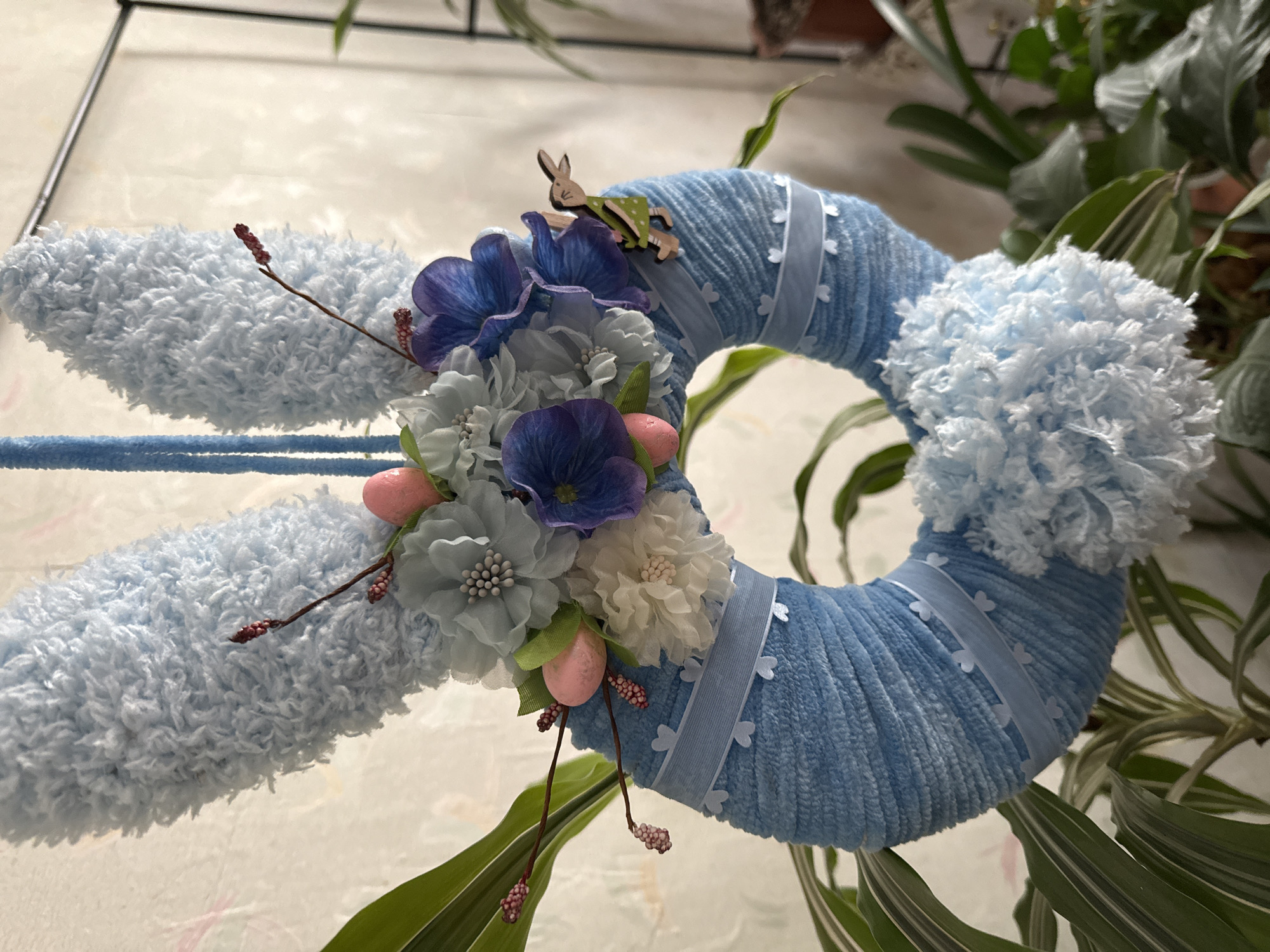 An Easter bunny decoration hangs in front of a light-colored, textured wall. The decoration is shaped like a bunny head with two fuzzy light blue ears extending upwards. A circular wreath, also covered in light blue yarn, is attached to the bottom of the ears. The wreath is adorned with various fabric flowers in shades of light blue, white, and purple, as well as small pink egg-shaped decorations. A thin light blue ribbon with small heart cutouts is wrapped around the wreath. A small wooden bunny figure, painted green and white, is attached to the side of the wreath. Below the wreath, a fluffy white pom-pom is attached. The decoration is surrounded by lush green leaves of indoor plants. The background suggests an indoor setting with a hint of a dark metal frame in the upper right corner.