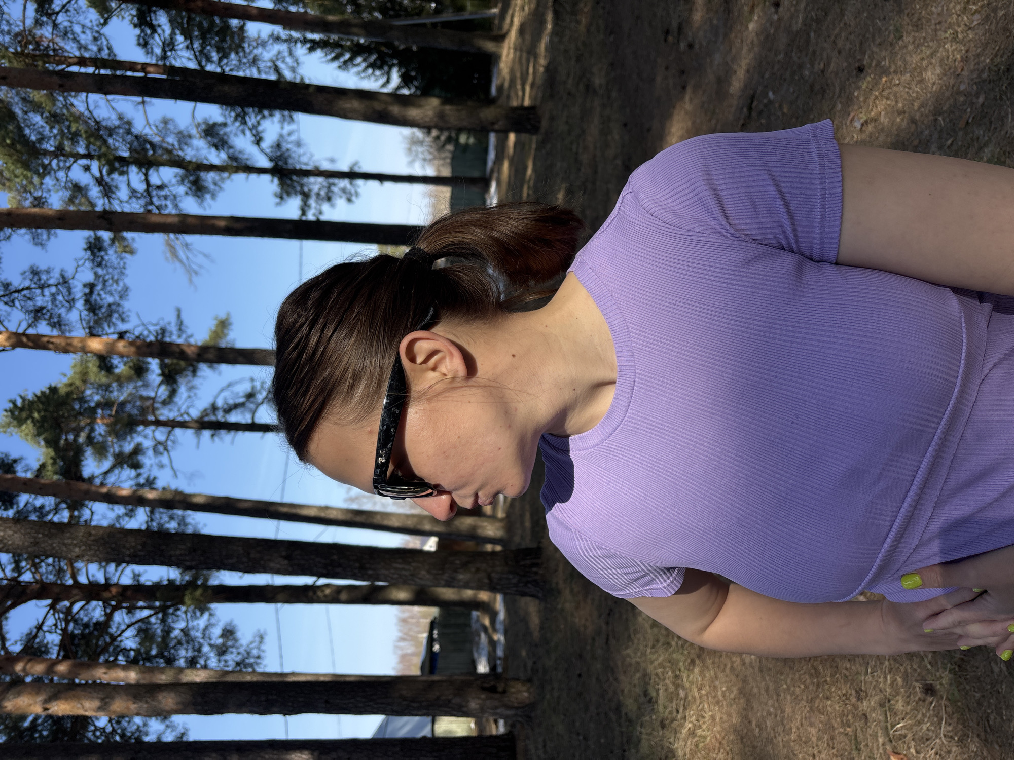 The scene depicts a female individual outdoors in the city of Раздолье, Russia. She is positioned in profile, facing left, captured from the mid-torso upwards. Her dark brown hair is pulled into a ponytail, and she wears black sunglasses with "Ray-Ban" branding visible on the right temple. Her attire is a short-sleeved, ribbed, light purple top. Her left hand, featuring neon green nail polish, rests on her left hip. The environment is characterized by numerous tall pine trees with exposed reddish-brown trunks and green canopies against a clear blue sky. The ground consists of dry, brown foliage and patches of melting snow. A residential structure with a dark roof and light-colored walls is partially visible in the distant left background, alongside a dark fence.