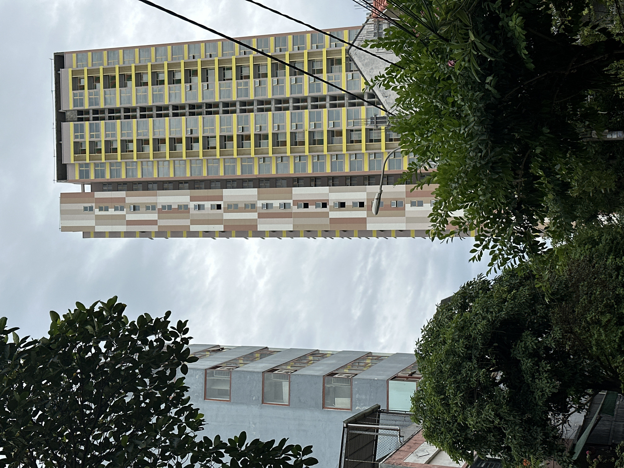 The image shows a view of two tall buildings in Đà Nẵng, Vietnam, under a cloudy sky. The building on the left is a light blue, multi-story structure with several visible windows. To its right, a taller building features a yellow facade with many windows framed in yellow. A narrower section of this building has a vertical pattern of beige and pink rectangular panels. In the foreground, lush green trees obscure the lower portions of the buildings. Power lines are visible in the upper right quadrant of the image. There are no people or animals visible. The scene appears to be an urban streetscape.