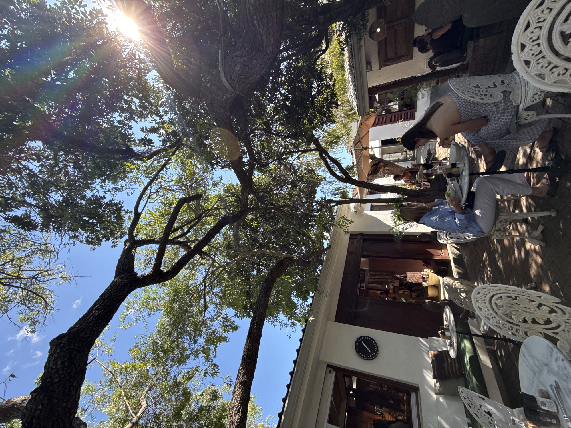 This image shows an outdoor café or restaurant in Cape Town, South Africa, as evidenced by the sign "at The Stranger's Club" on the building. The scene is oriented towards an outdoor seating area under trees, with a bright sun shining through the leaves. Several people are seated at white, ornate metal tables and chairs, appearing to be enjoying food and drinks. One person is looking down at a table, while another person is reading a document. The café is situated in front of a white building with dark-framed windows and doors. The environment is sunny and seems relaxed, suggesting a leisurely setting.
